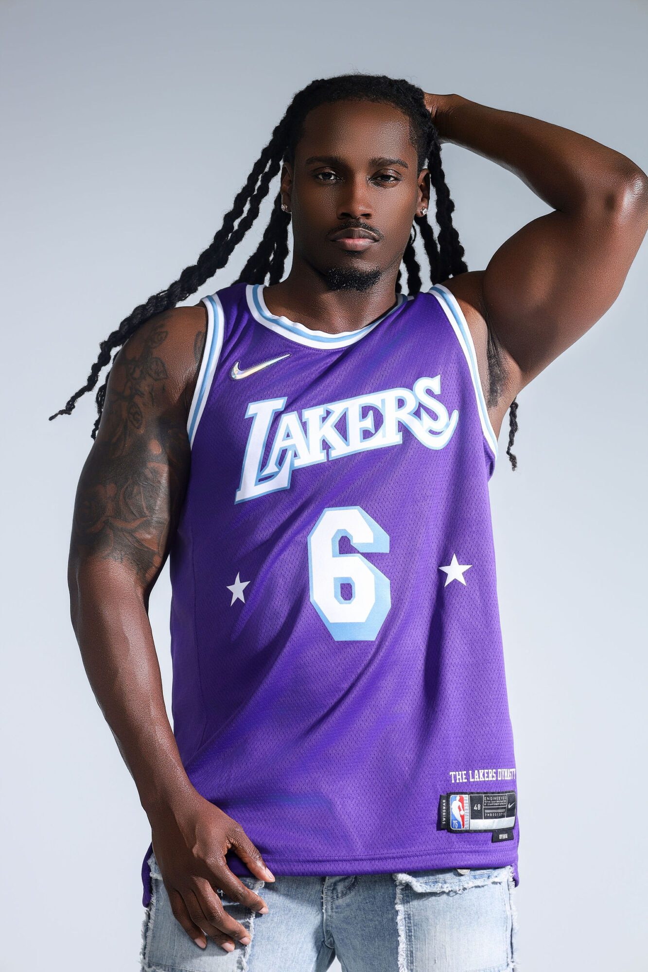 Man with long dreadlocks wearing a purple Los Angeles Lakers basketball jersey, standing with one hand behind his head.