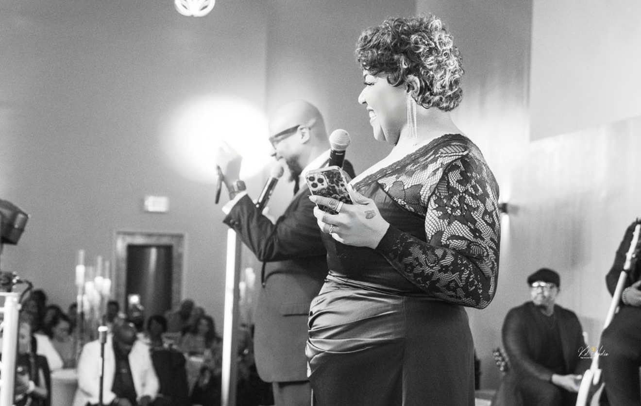 Two people on stage, one holding a microphone and the other holding a phone, with an audience in the background.