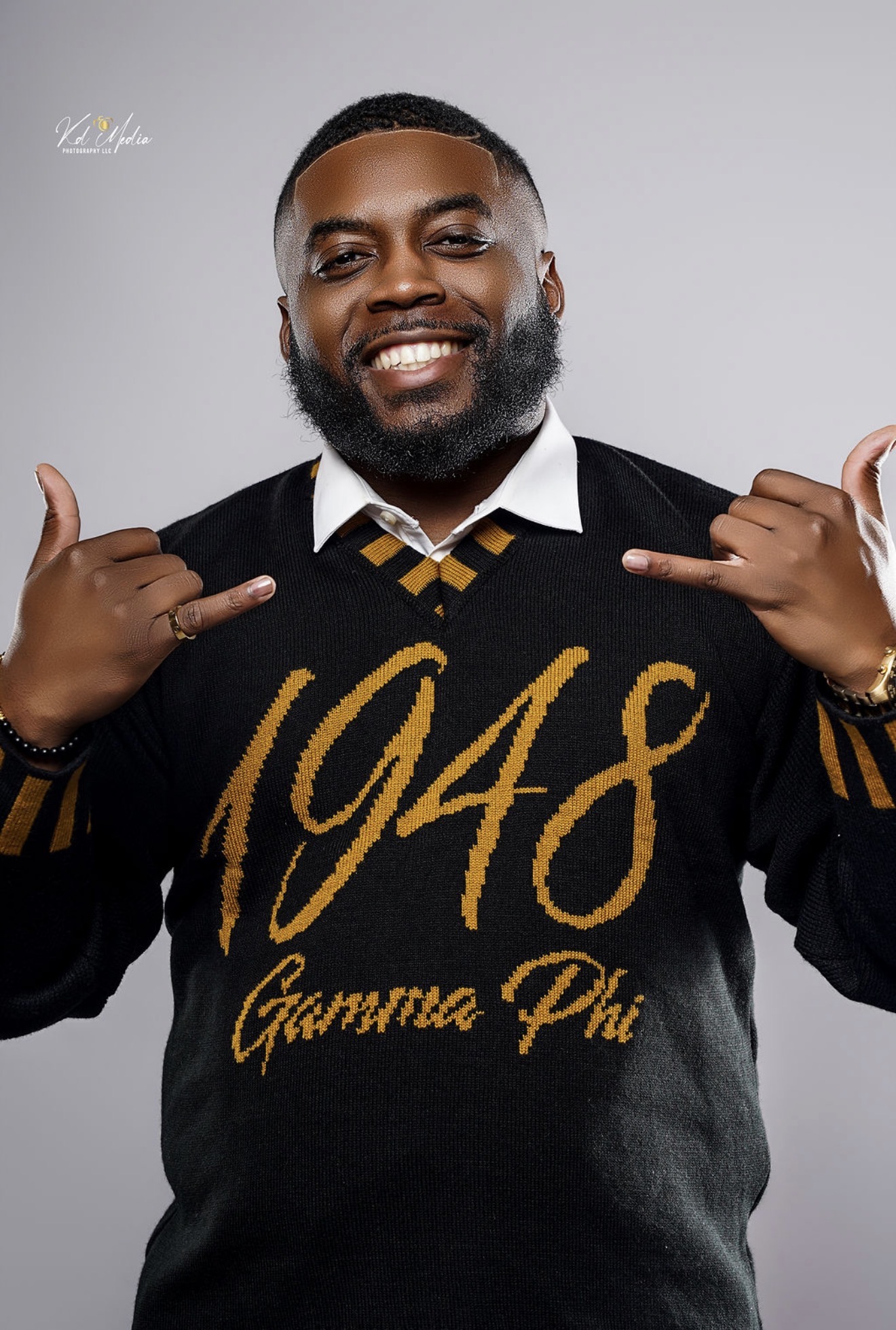 Man with a beard smiling, wearing a black sweater with gold text, making a shaka sign with both hands.