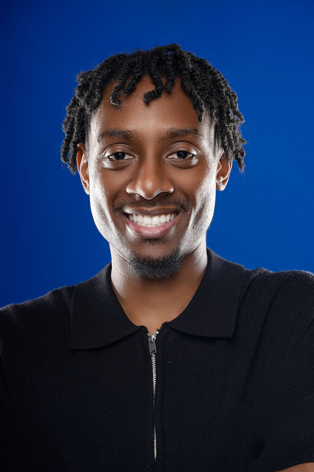 Smiling person with short curly hair, wearing a black zip-up top, against a blue background.