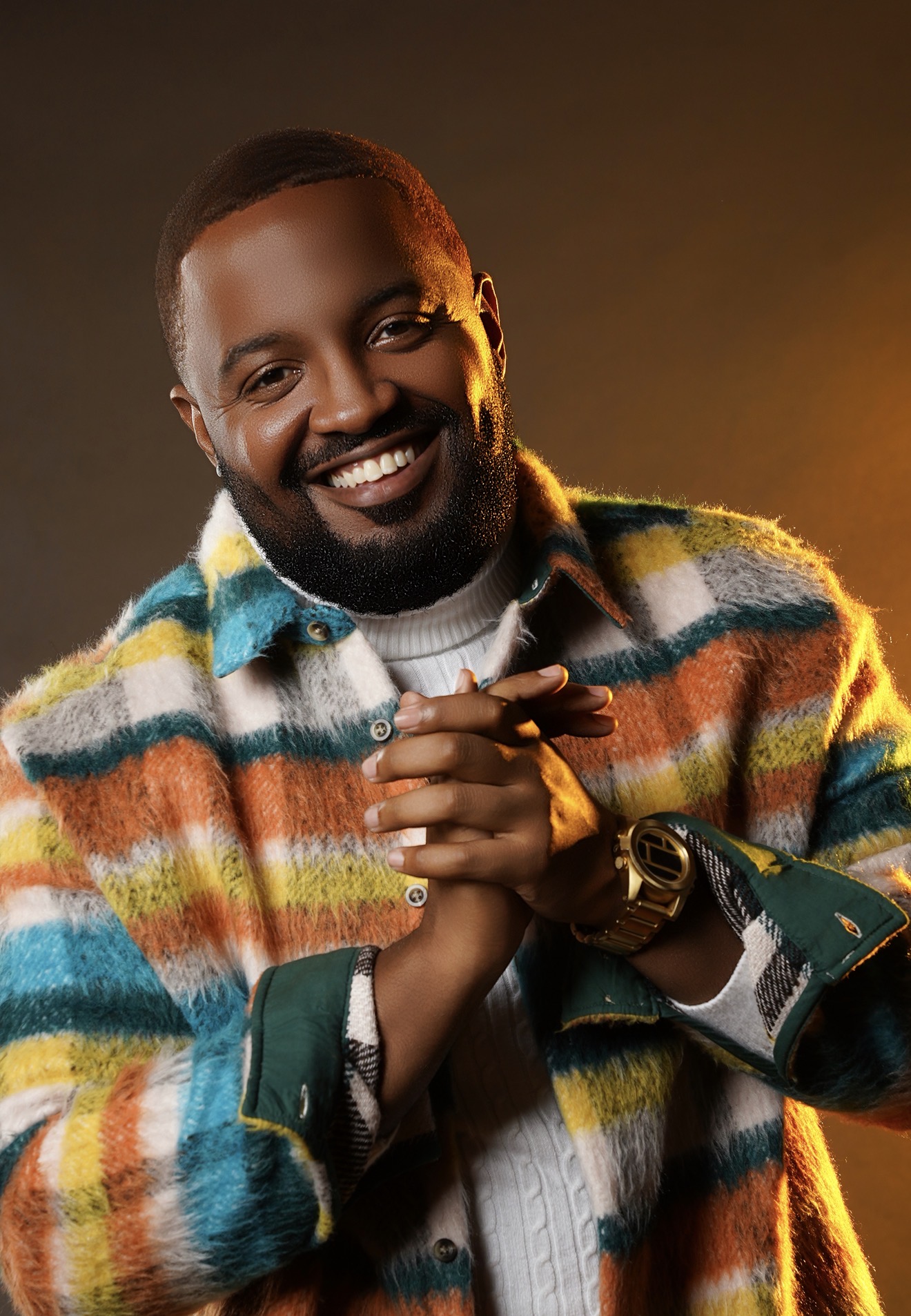 Smiling man with short hair and beard, wearing a colorful striped jacket, clasping hands, against warm background.
