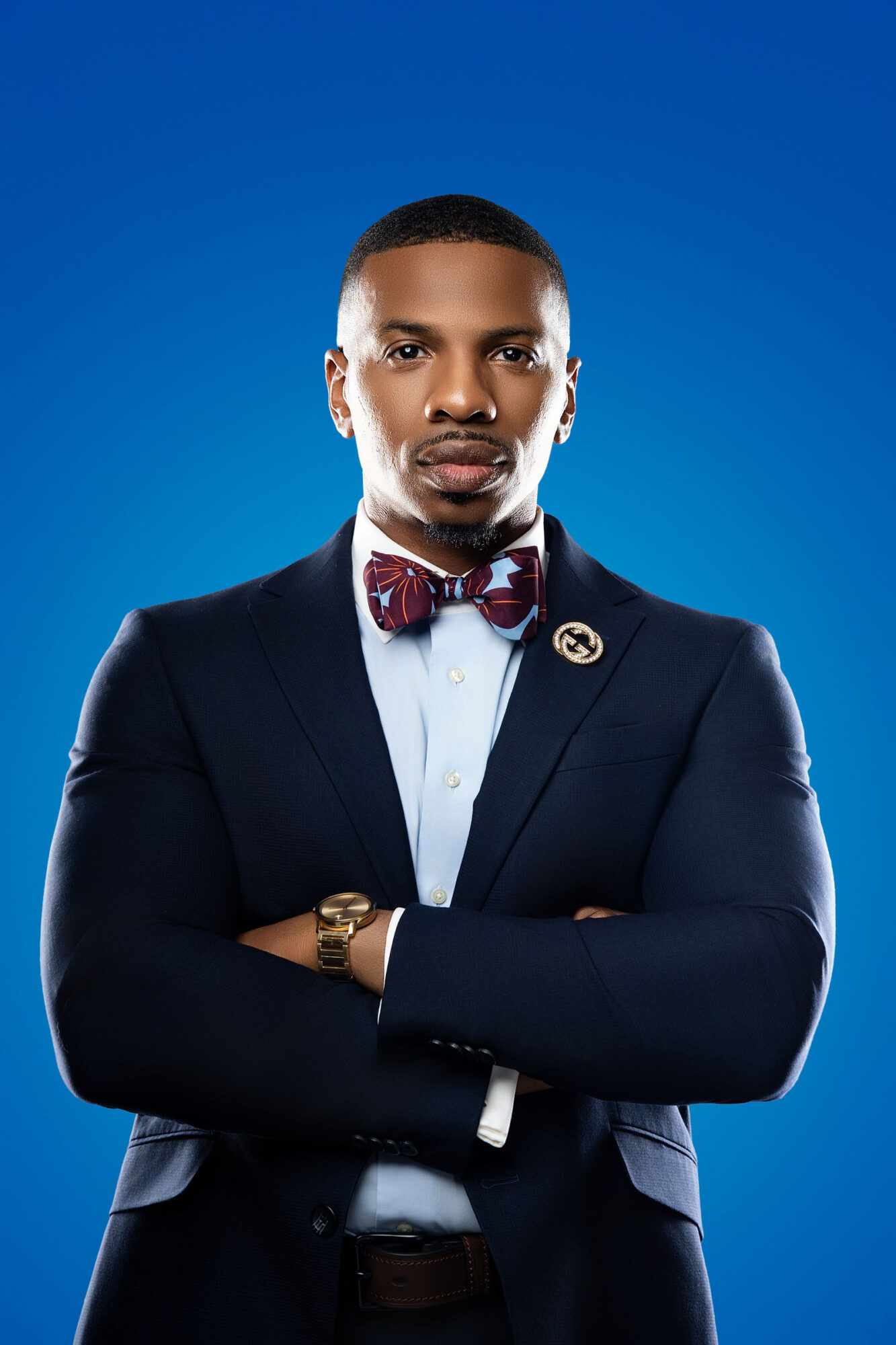 Man in formal suit with bow tie and watch, arms crossed, against blue background.