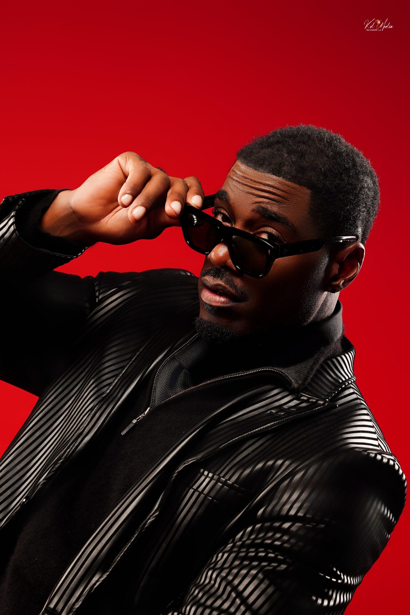 Man with short hair and sunglasses adjusting glasses against a red background