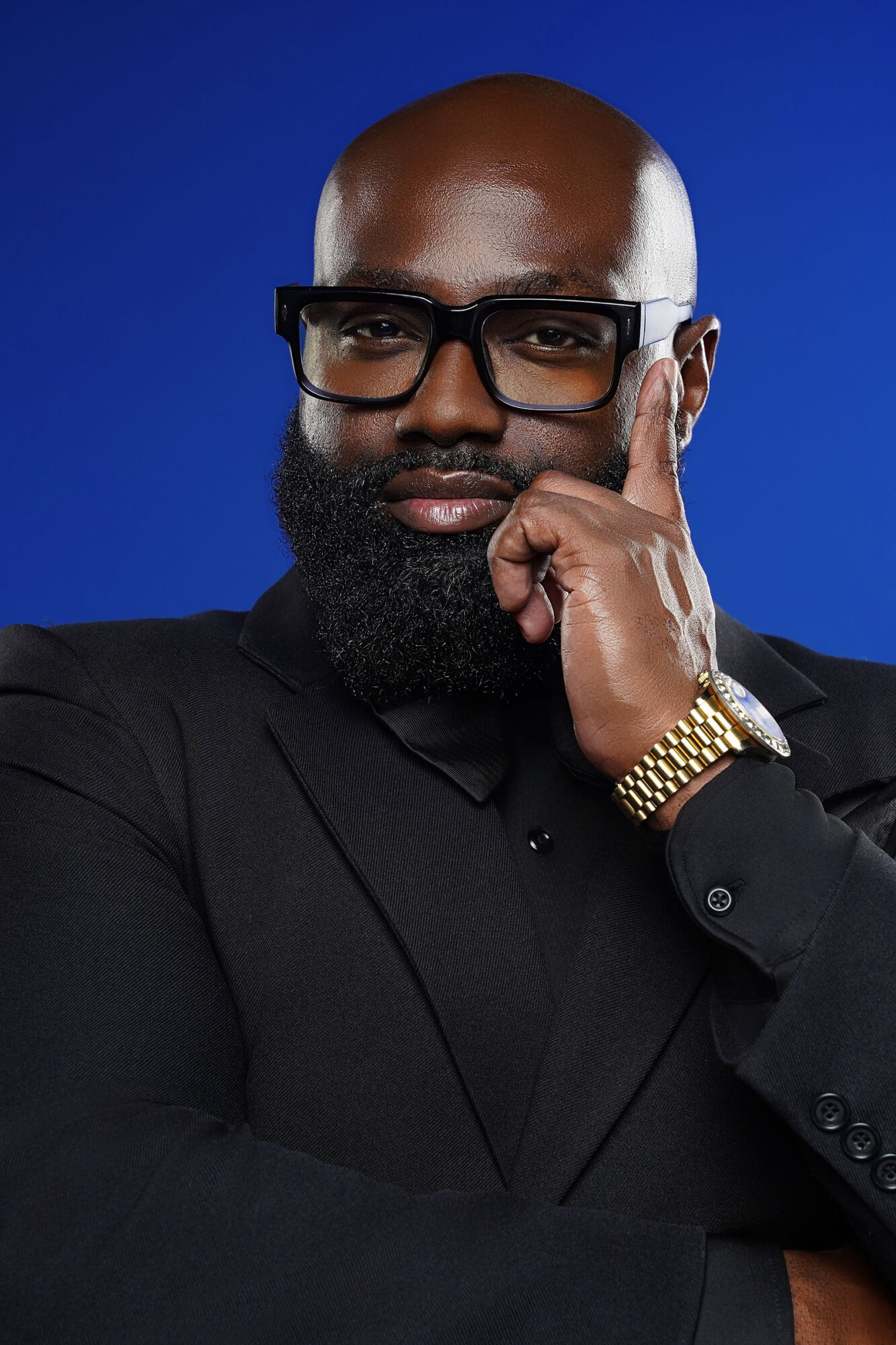 Man with glasses and a beard, wearing a black suit, touching his face with his right hand, against a blue background.