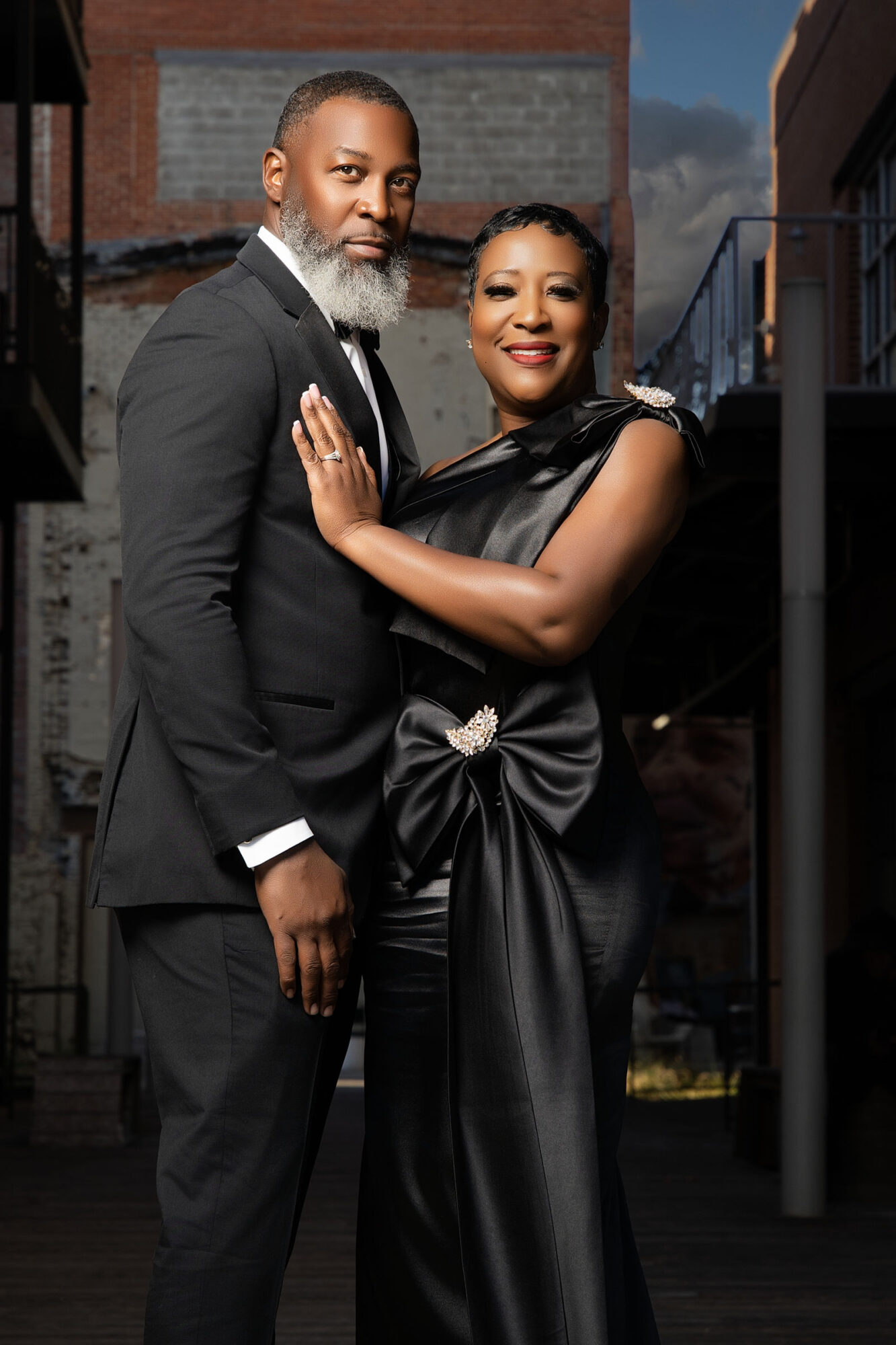 A well-dressed couple standing outdoors, smiling, with buildings in the background, during evening or dusk.