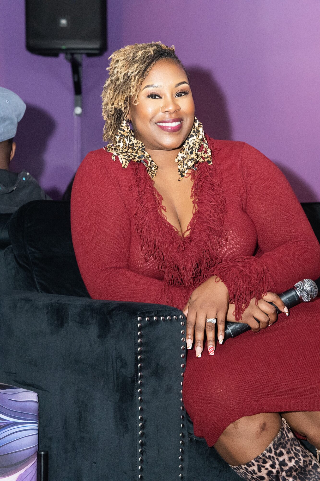 Woman with curly blonde hair smiling, wearing a red dress, holding a microphone, sitting on a black chair against a purple background.