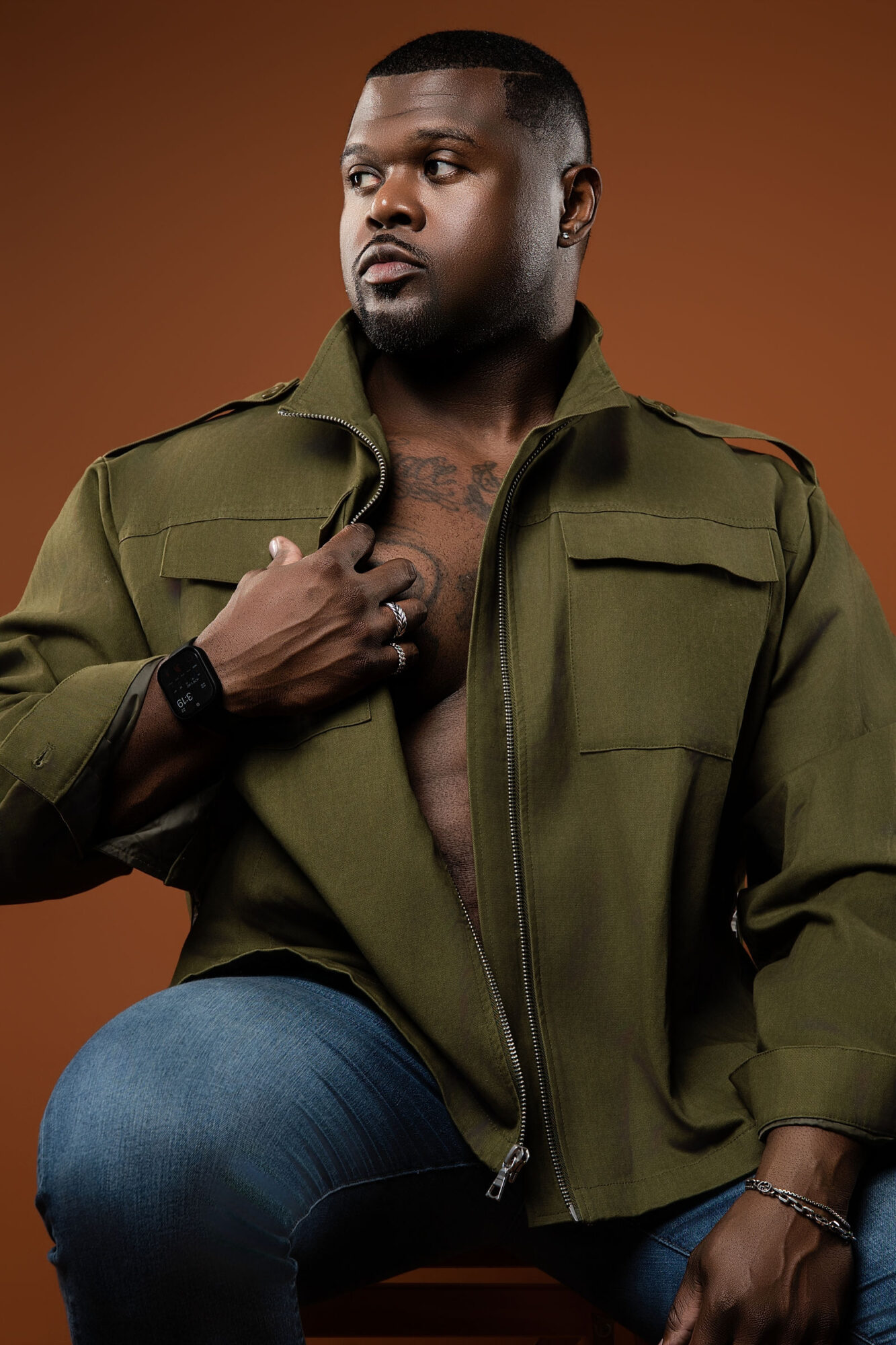 Man with short hair and beard wearing an open military-style jacket, sitting against a brown background.