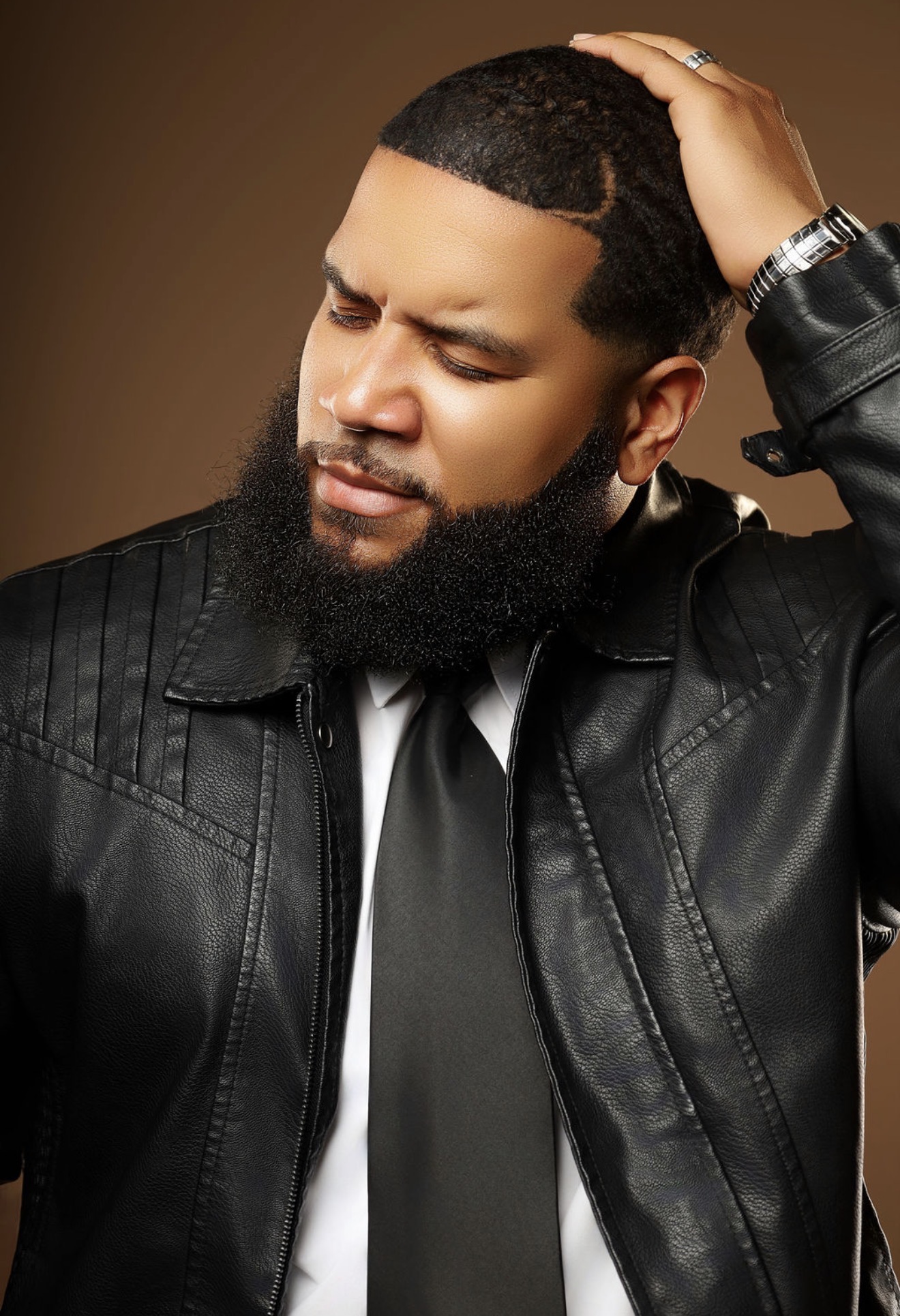 Man with a beard and short hair, wearing a black leather jacket and tie, touching his head with a neutral expression.