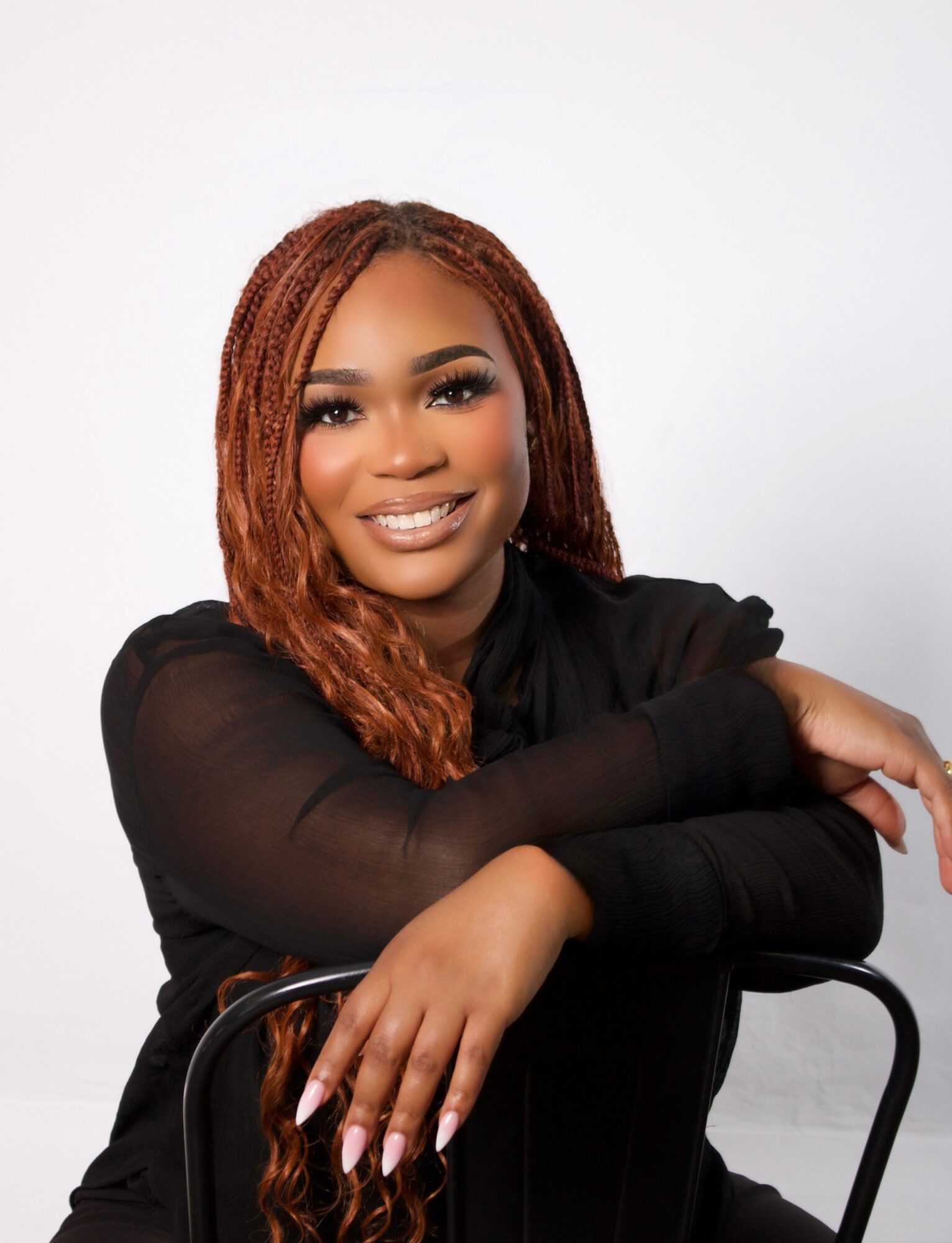 Smiling woman with long, reddish-brown braided hair, wearing a black top, resting her arms on a chair, against a white background.