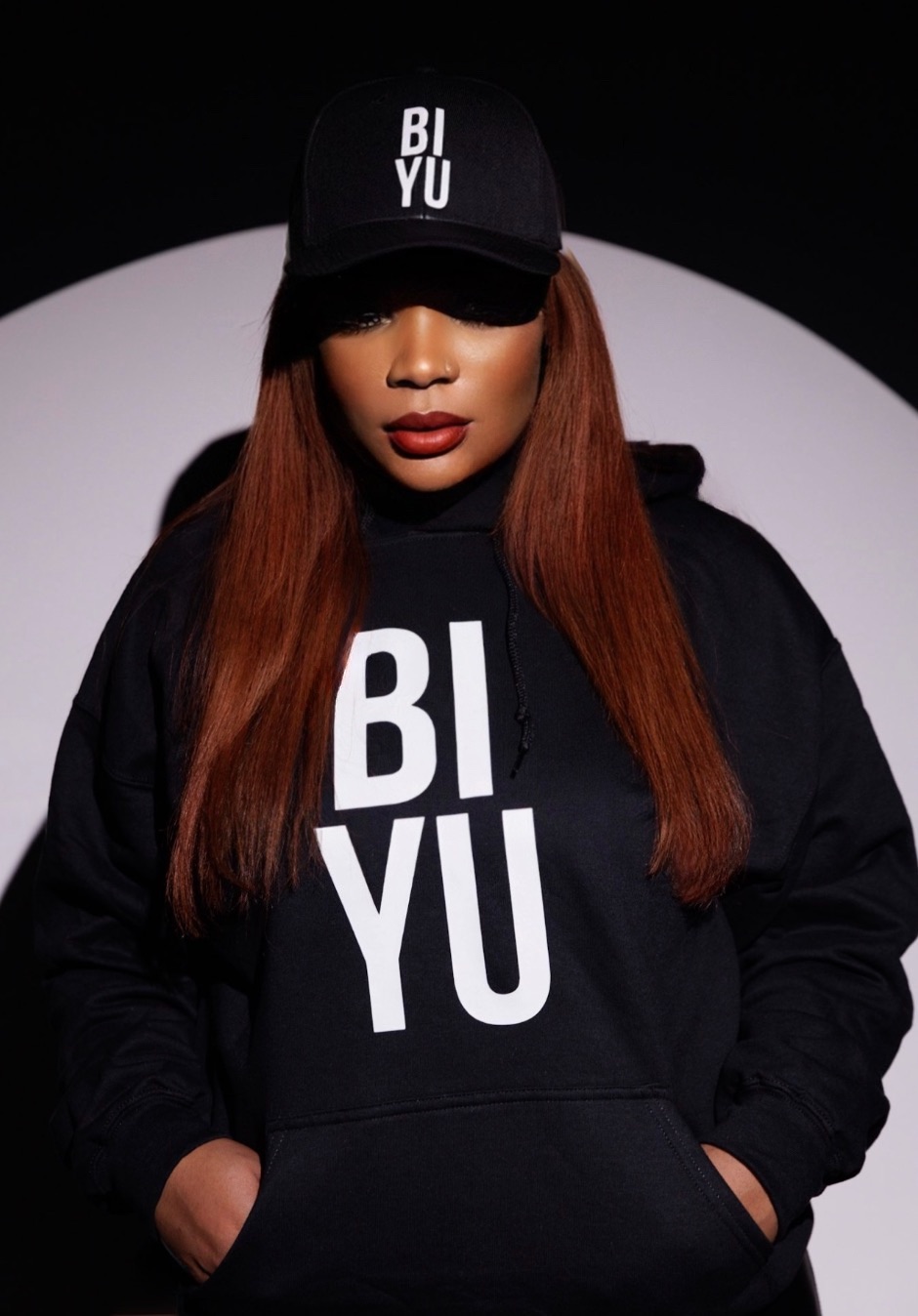Woman wearing a black cap and hoodie with white text, standing against a dark background.