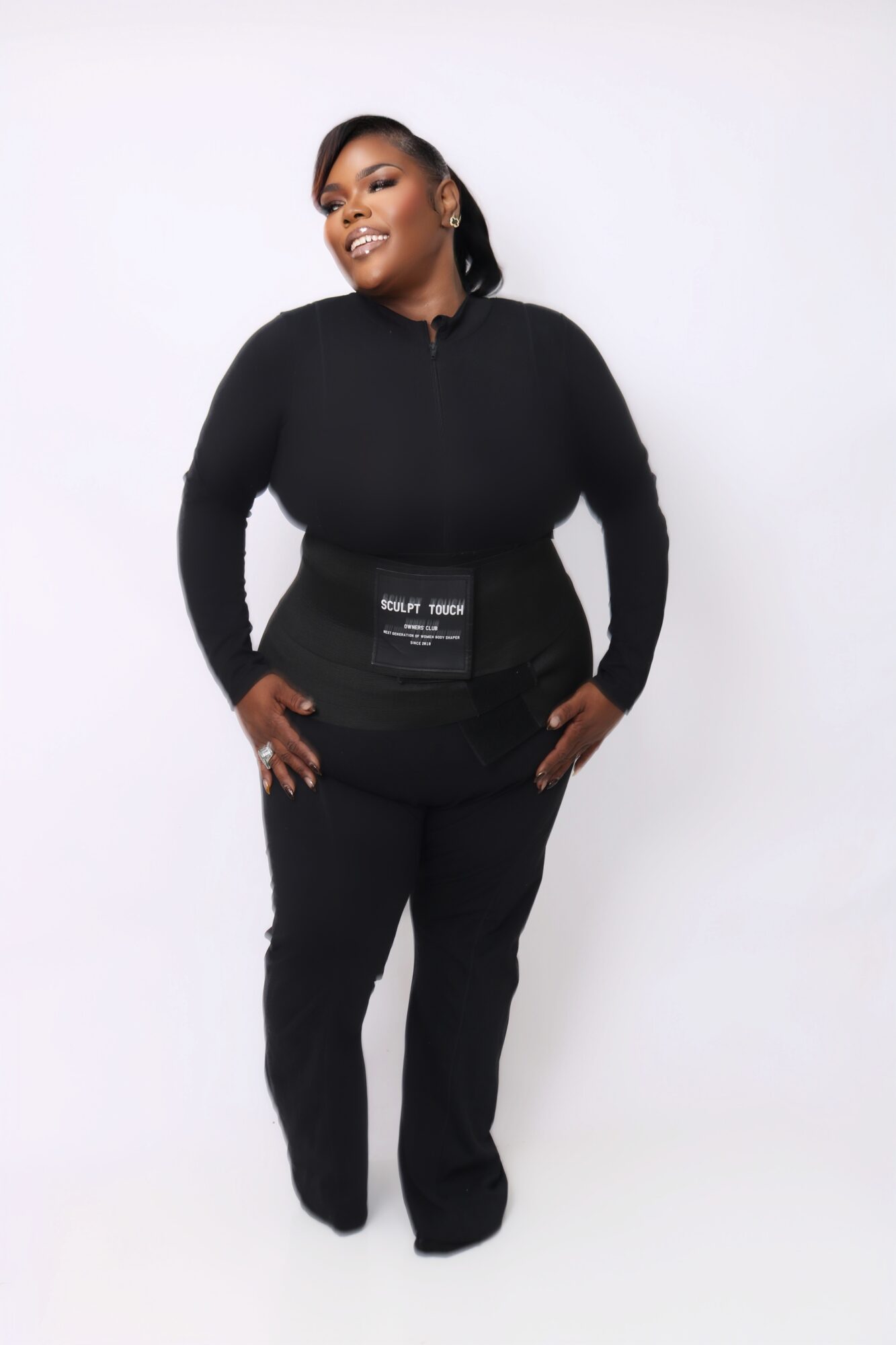 A woman with dark hair and earrings wears a black zip-up top and black pants, smiling with hands on hips.