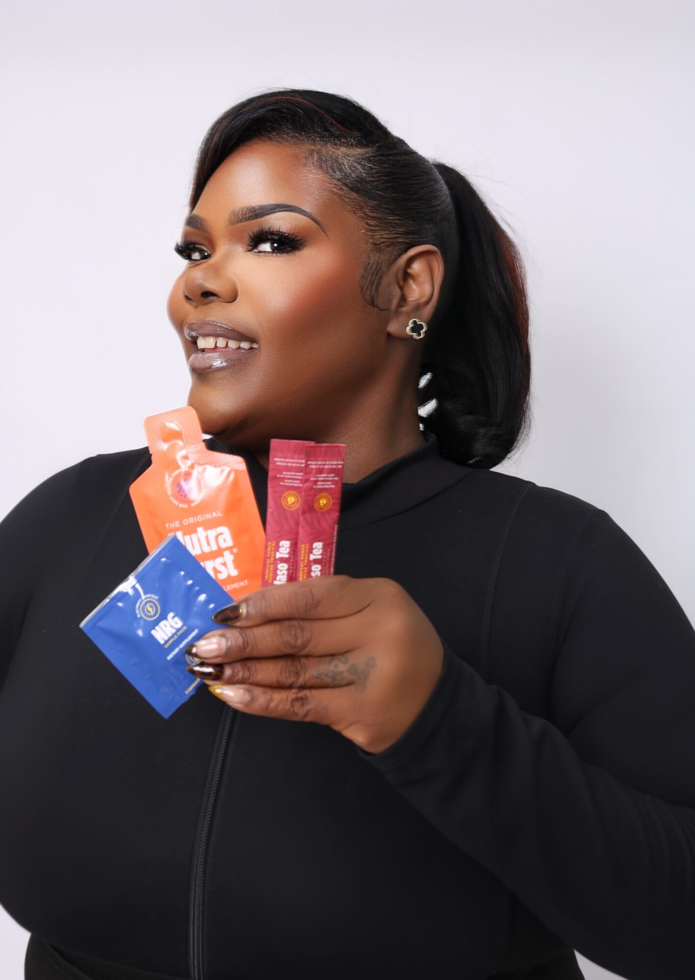 Woman with dark hair in a ponytail holding colorful snack packages, smiling, wearing a black top, against a plain background.