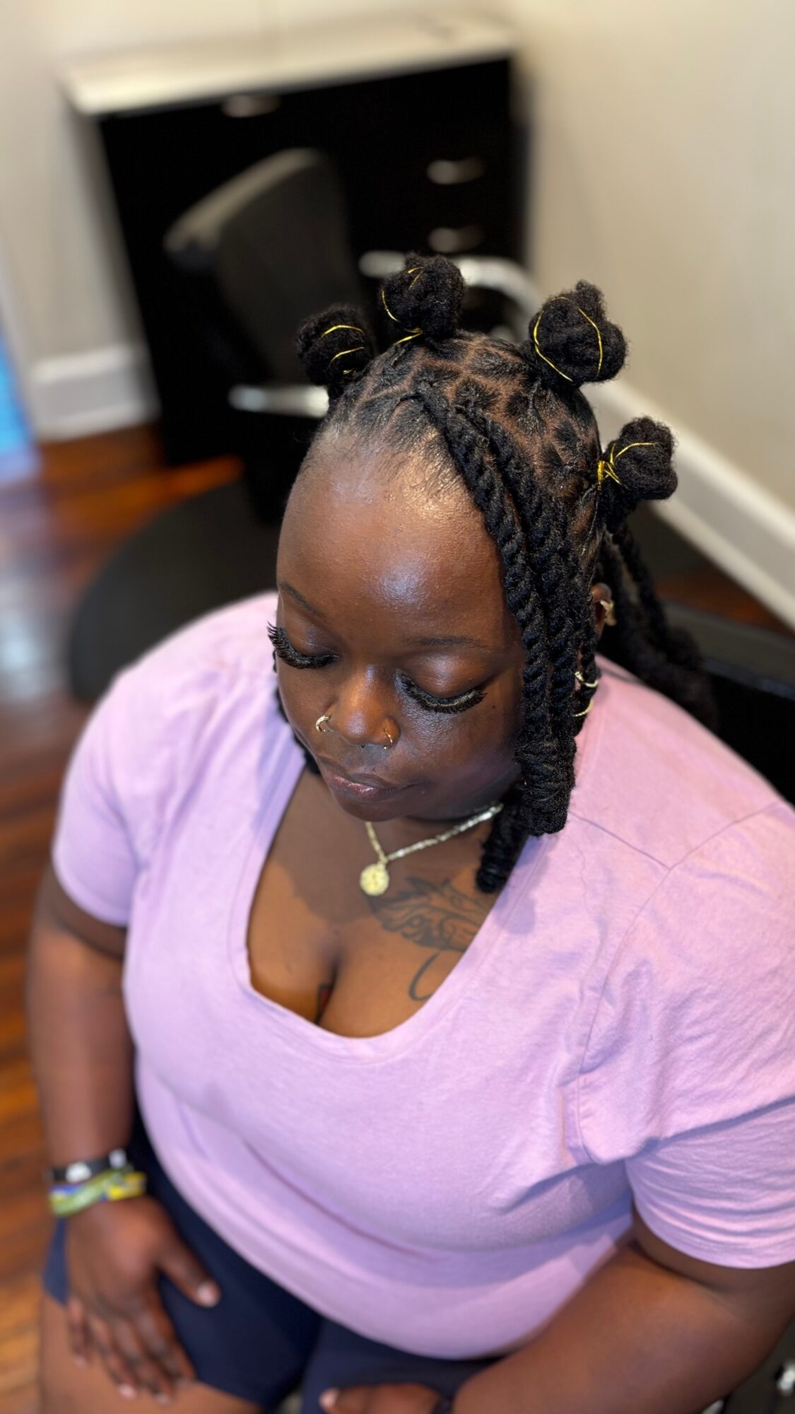 Woman with braided hairstyle and bantu knots, wearing a purple top, sitting indoors with a black chair and furniture in background.