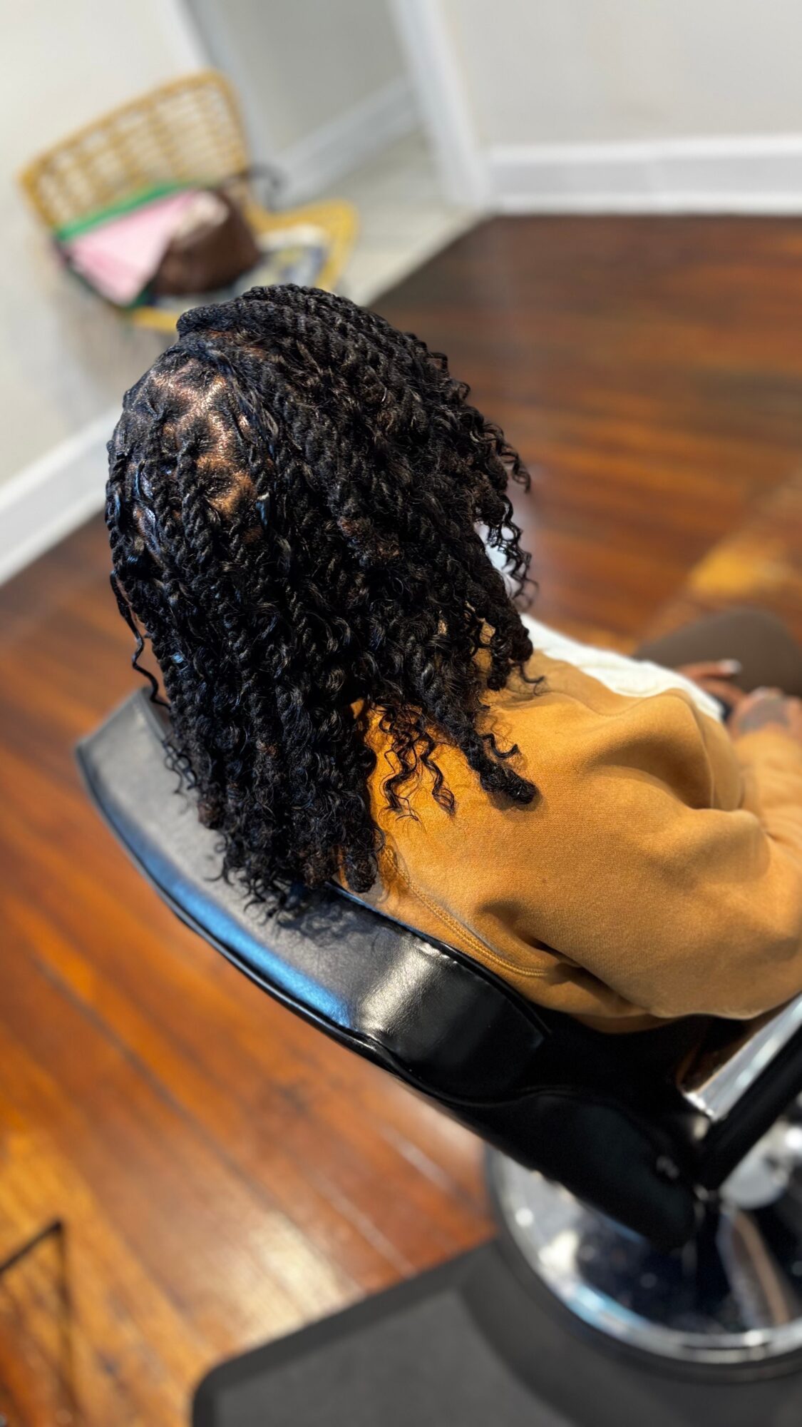 Person with curly hair sitting in a chair indoors, with a basket and a pink item in the background.