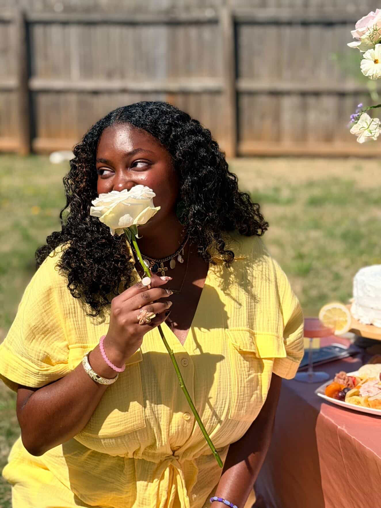 Woman in yellow dress holding a white flower near her face outdoors, with a wooden fence in background.