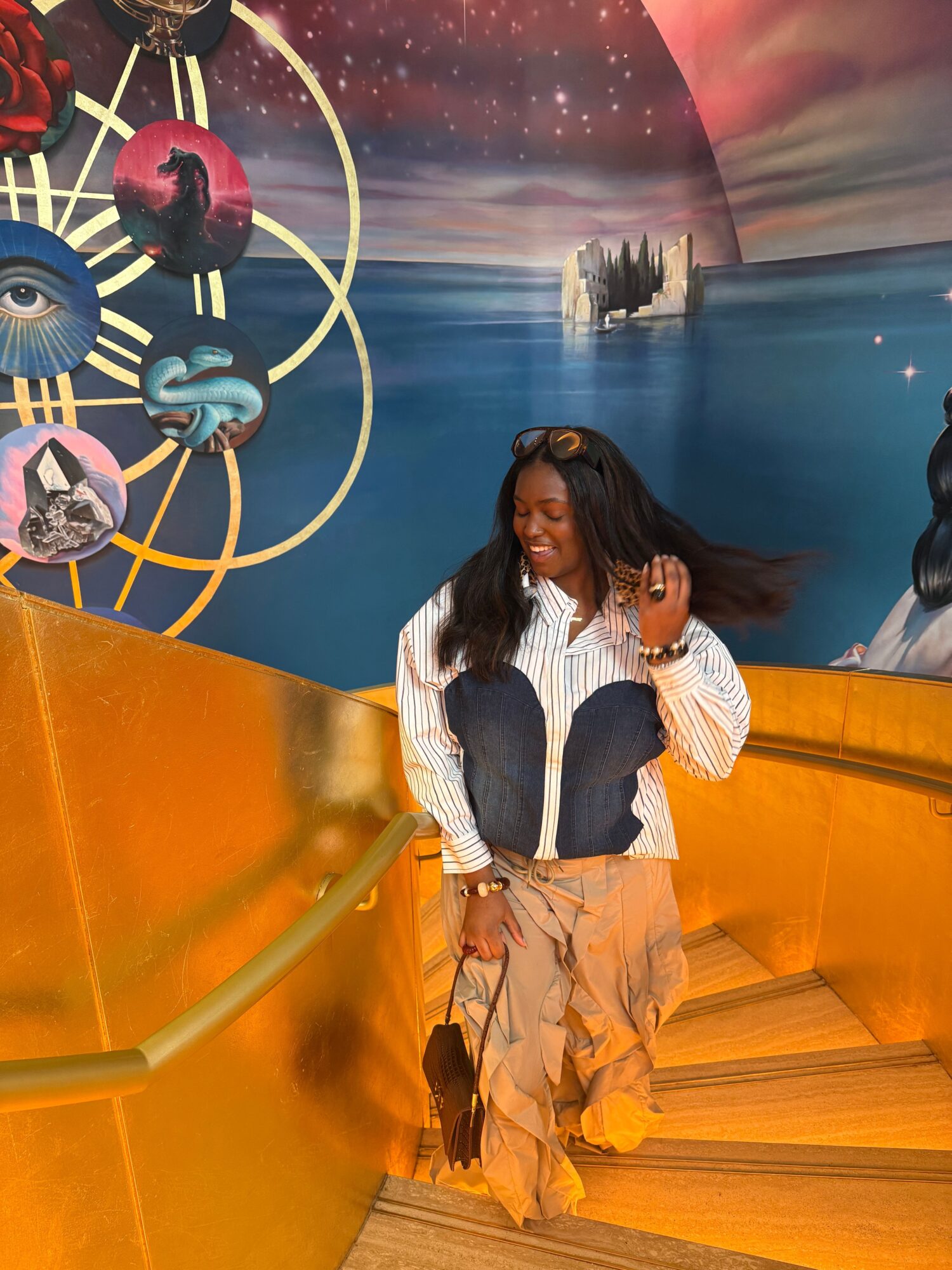 Woman with long dark hair, wearing a striped shirt and beige pants, standing indoors near a colorful backdrop with a yellow railing.