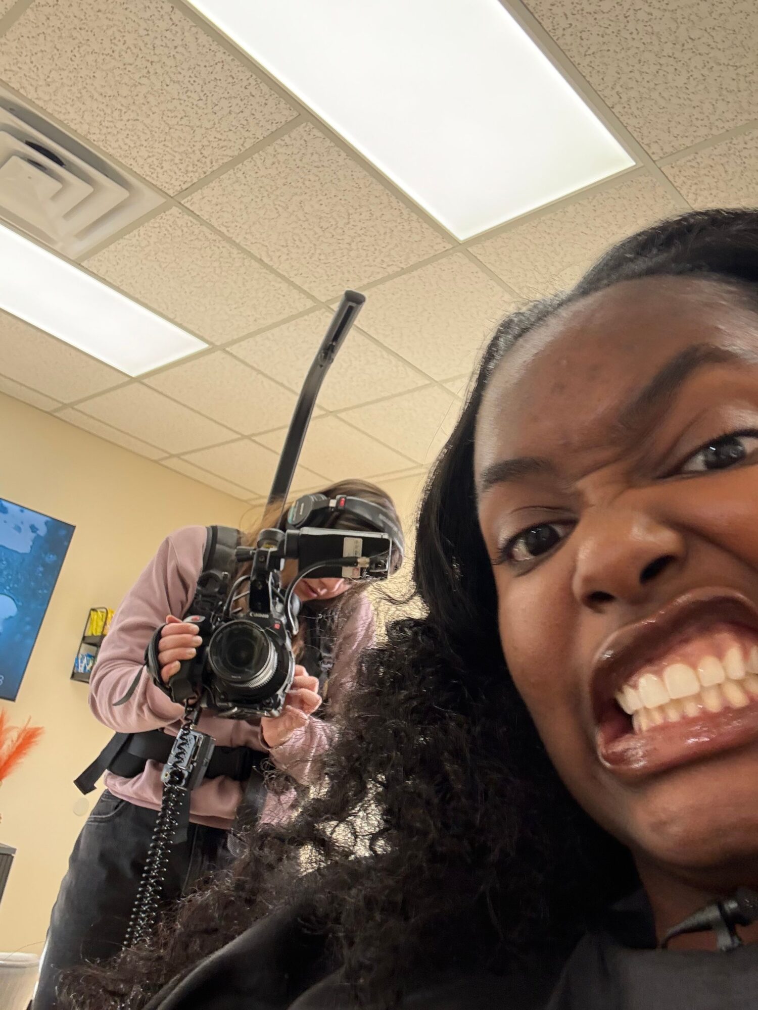 Person smiling and taking a selfie indoors with ceiling tiles and a camera in the background.