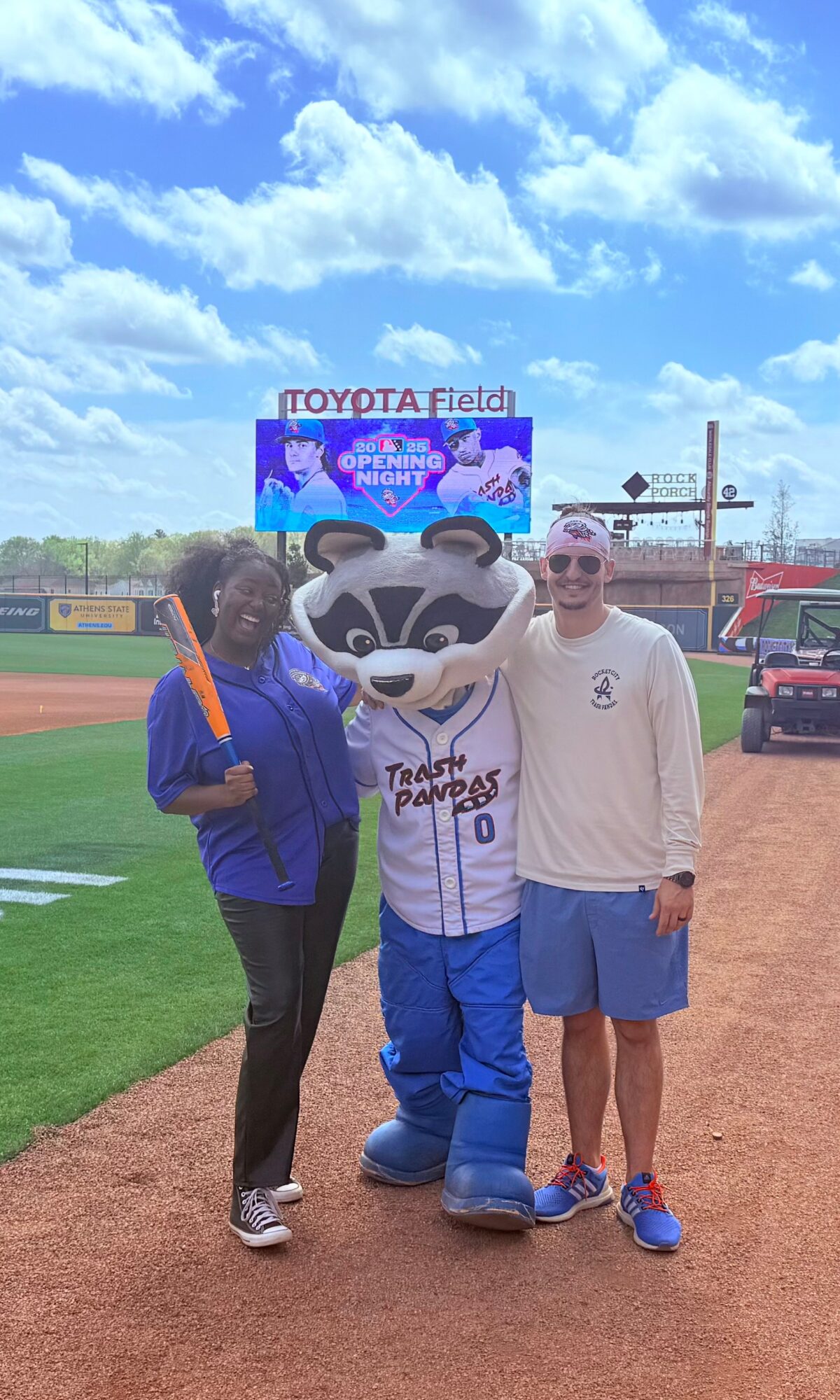 Two people and a mascot pose on a baseball field with a scoreboard and blue sky in background.