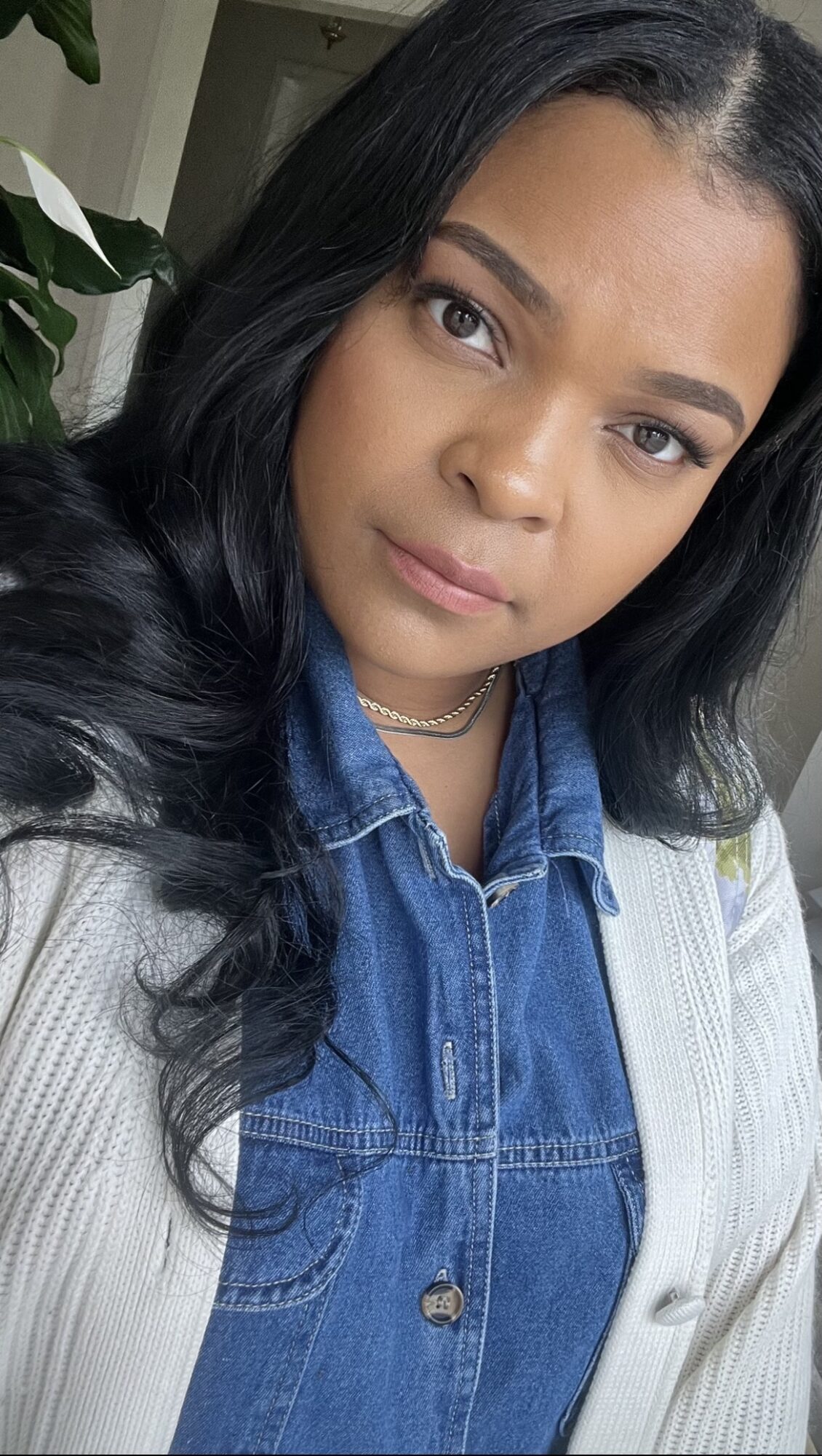 Young woman with long black hair, wearing a denim shirt and white cardigan, looking at the camera.