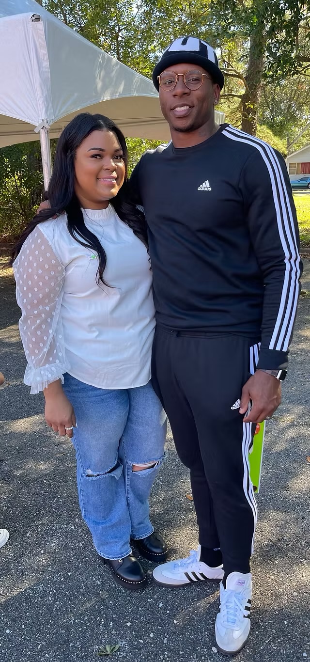 Two people standing outdoors, smiling, with trees and a tent in the background.