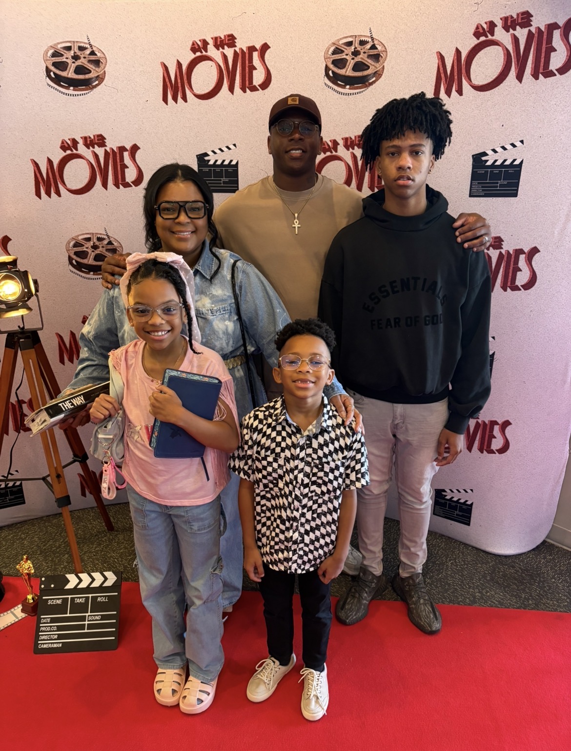 Group of five people standing on a red carpet in front of a backdrop with film-related graphics and text, smiling at the camera.