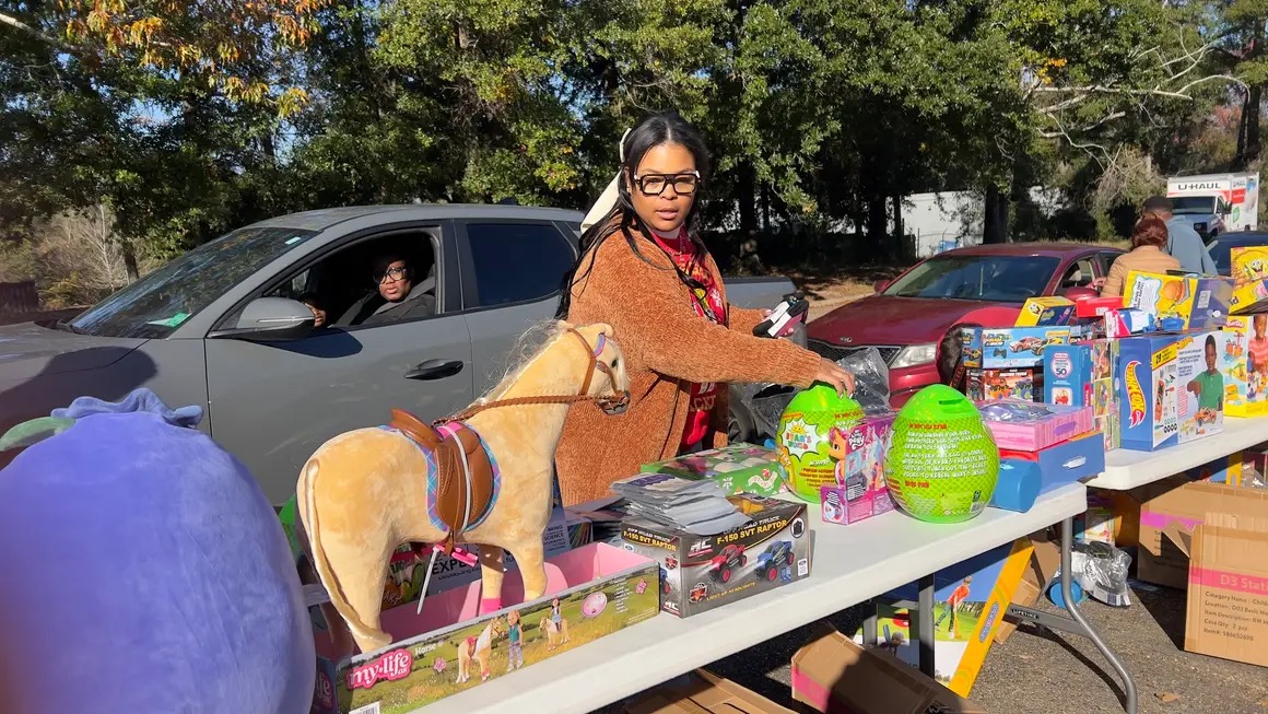 Woman with glasses and braids at outdoor table with toys and a dog, trees and cars in background.