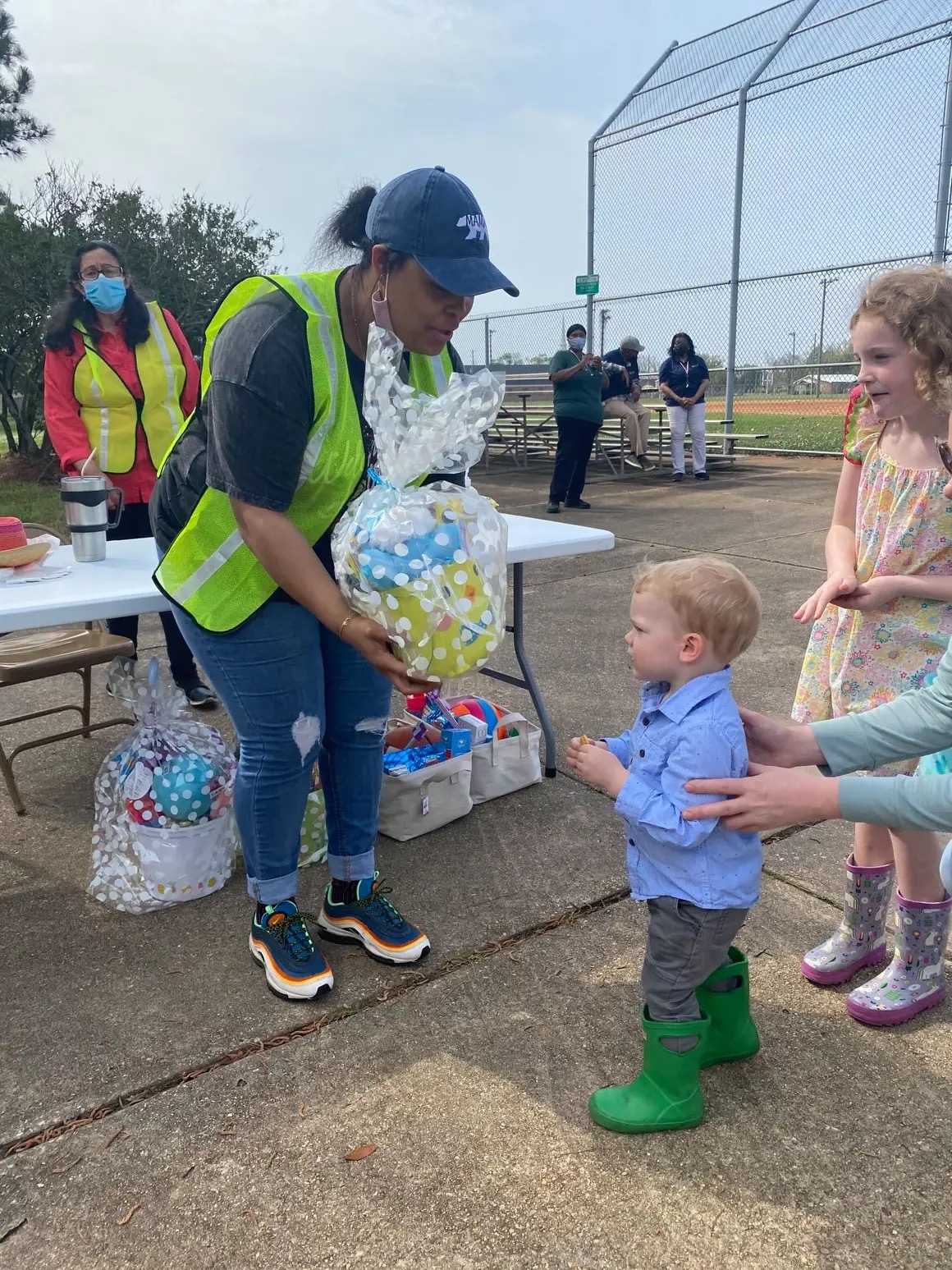 Woman in a yellow vest and blue cap hands a gift to a young boy in green rain boots at an outdoor event, with children and adults nearby.