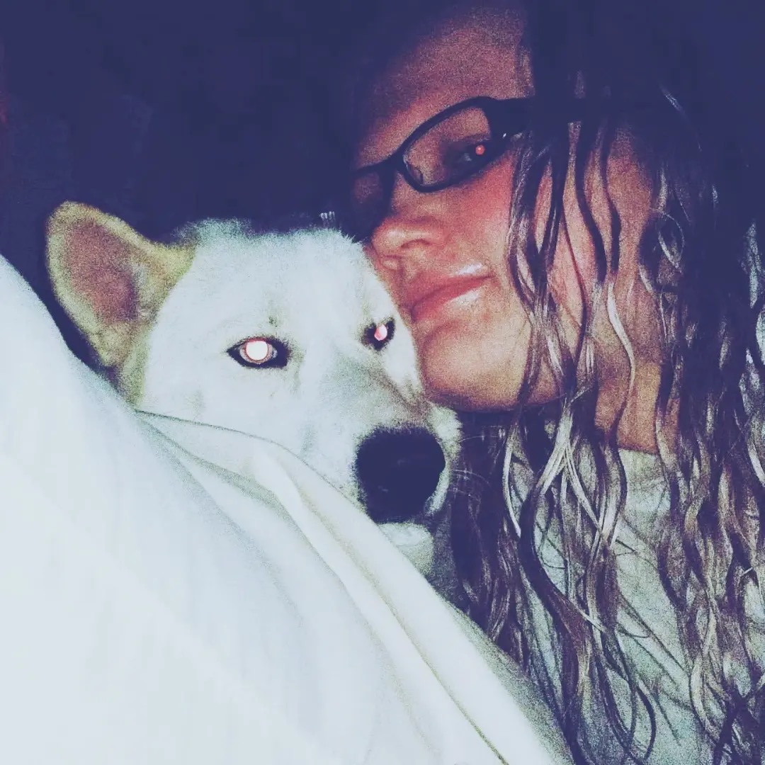 Woman with glasses and long hair close to a white dog with blue eyes, both resting on a white surface.