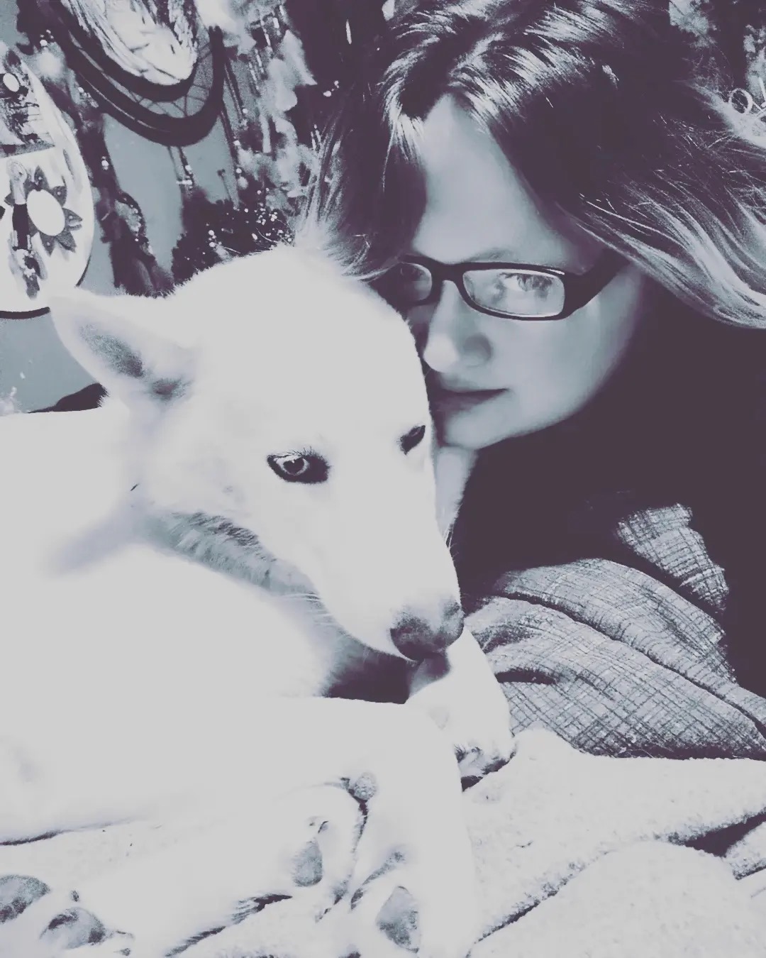 Person with glasses and long hair close to a white dog with blue eyes, indoors with patterned background.