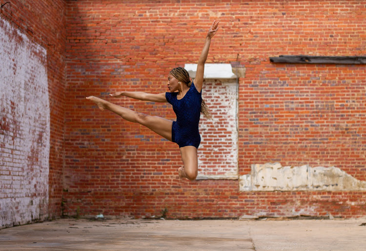 A young girl in a blue dress performs a leap against a brick wall, with arms and legs extended.