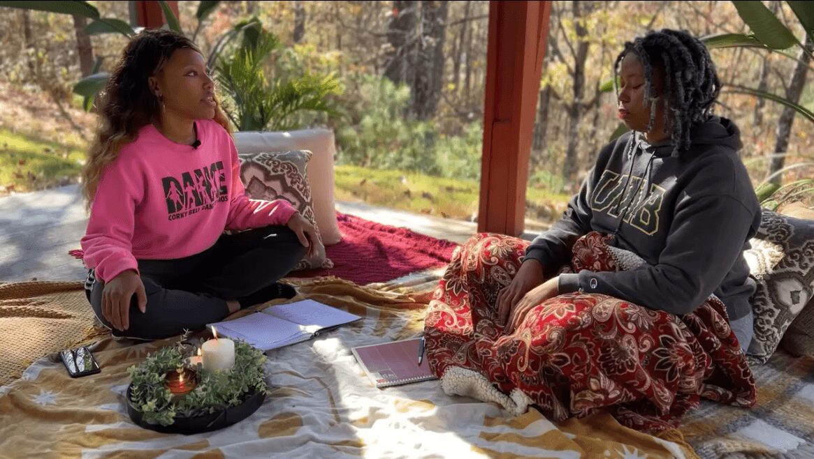 Two women sit on a blanket outdoors, engaged in conversation with trees and sunlight in the background.