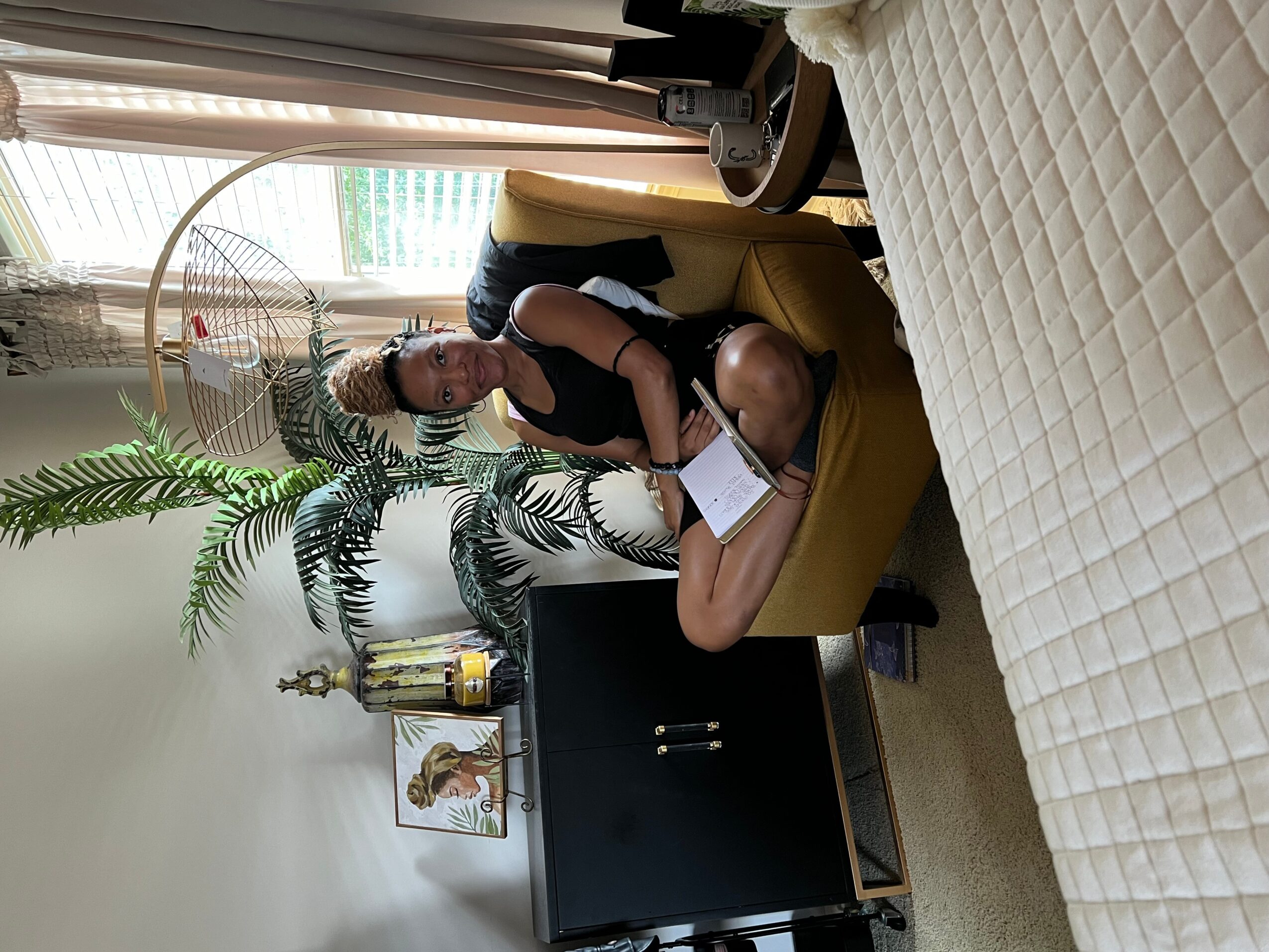Person sitting on a yellow chair holding a tablet, with a large plant and a trophy nearby, in a room with a bed.
