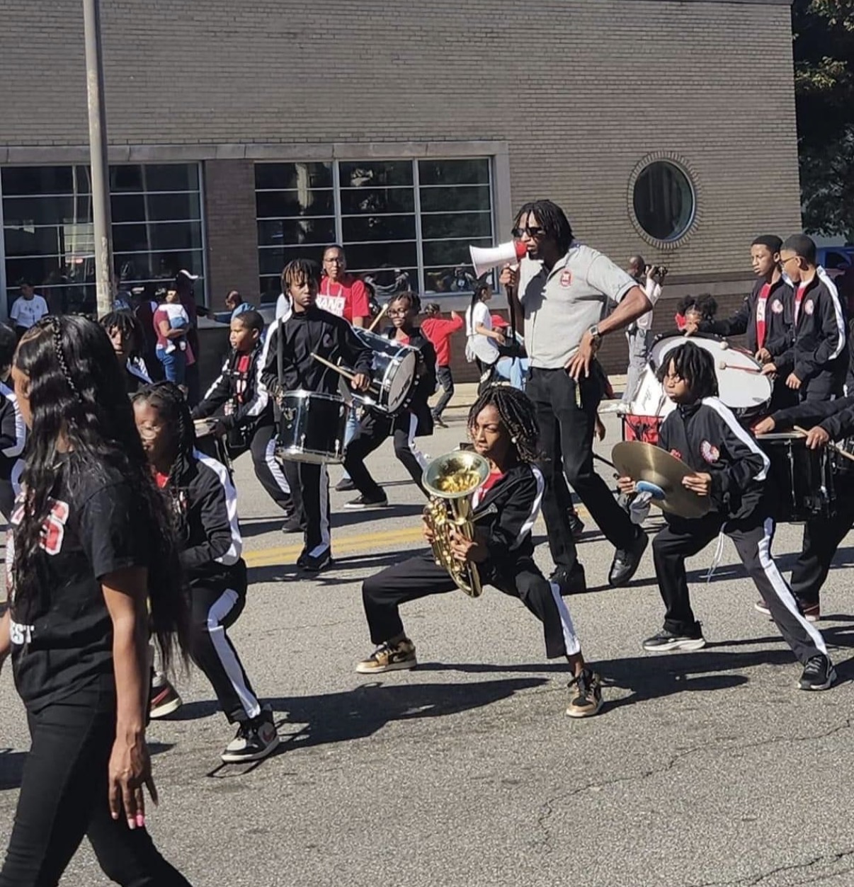 Group of children performing music outdoors, some playing drums and others holding instruments, with a woman speaking into a microphone.
