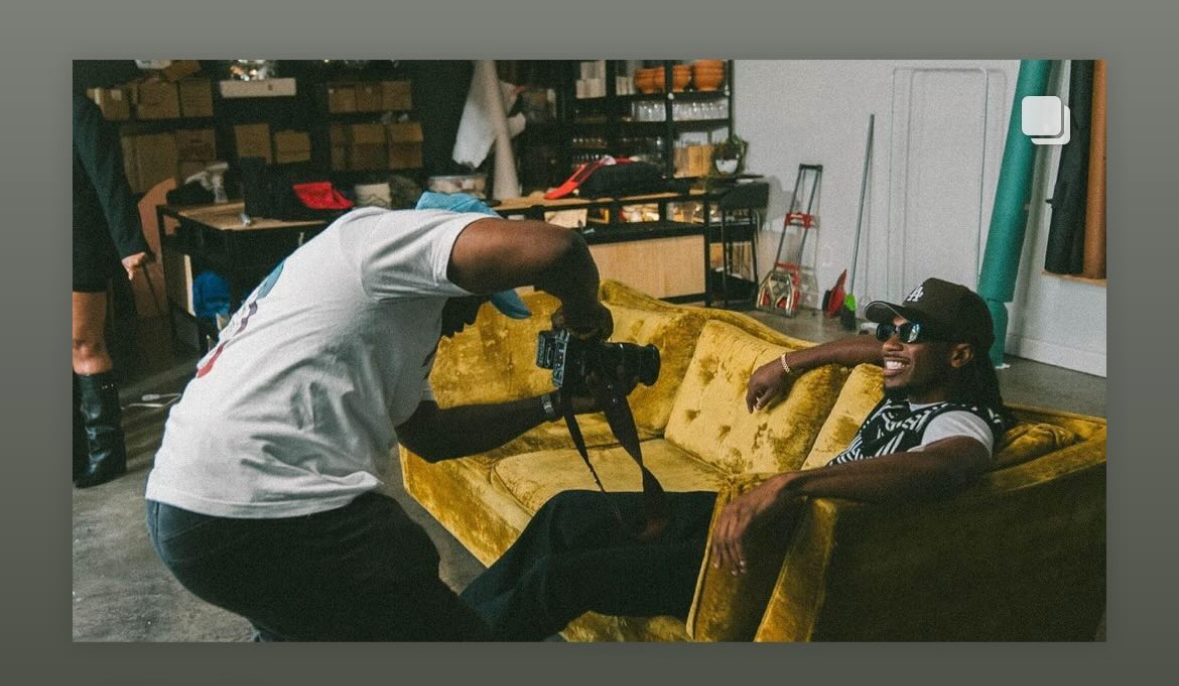 Person filming another person sitting on a yellow couch in a room with shelves and equipment, smiling and wearing sunglasses.