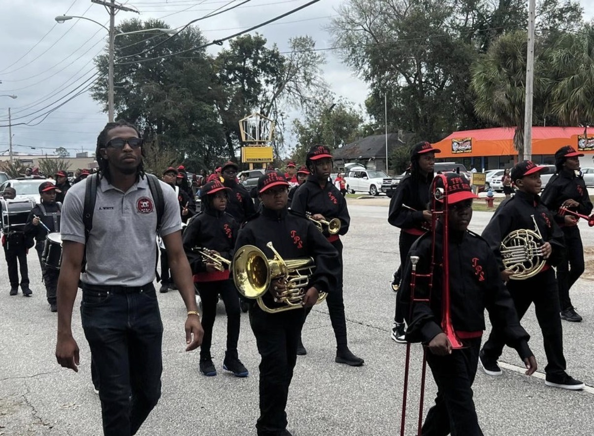 Group of people walking on a street, some holding musical instruments, with trees and buildings in the background.