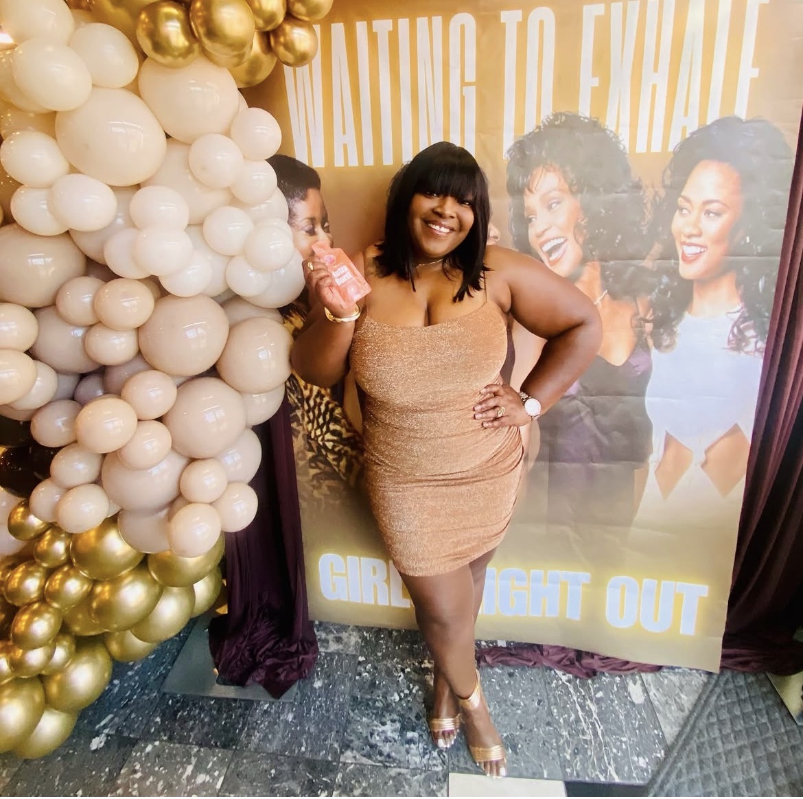 Woman in a beige dress smiling, holding a pink item, standing in front of a decorated backdrop with balloons and images of women.