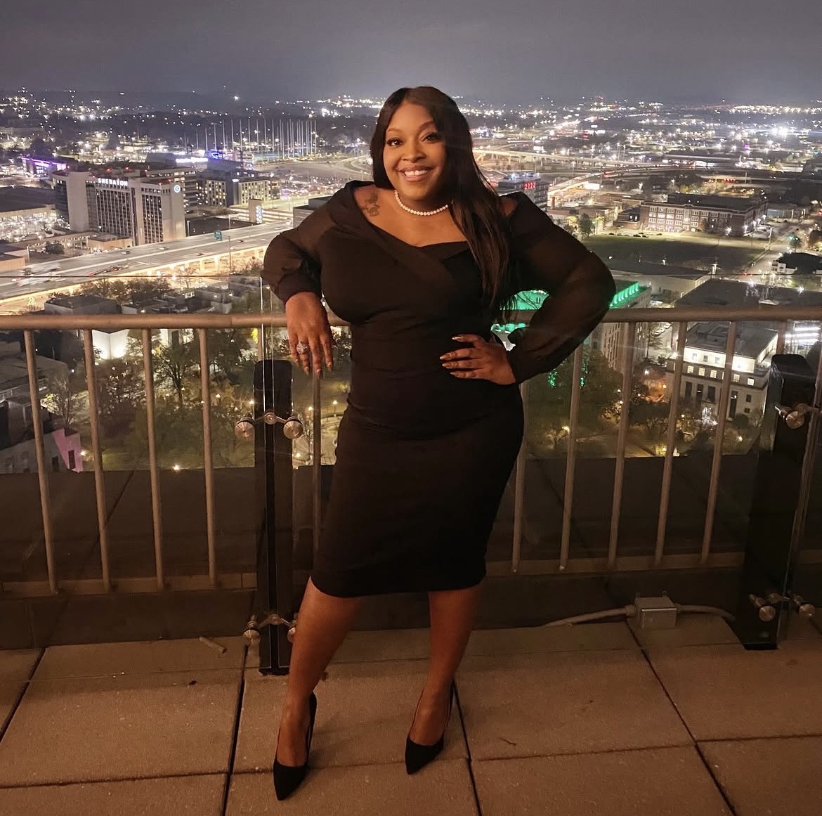 Woman in black dress and heels standing on balcony at night with city lights in background.