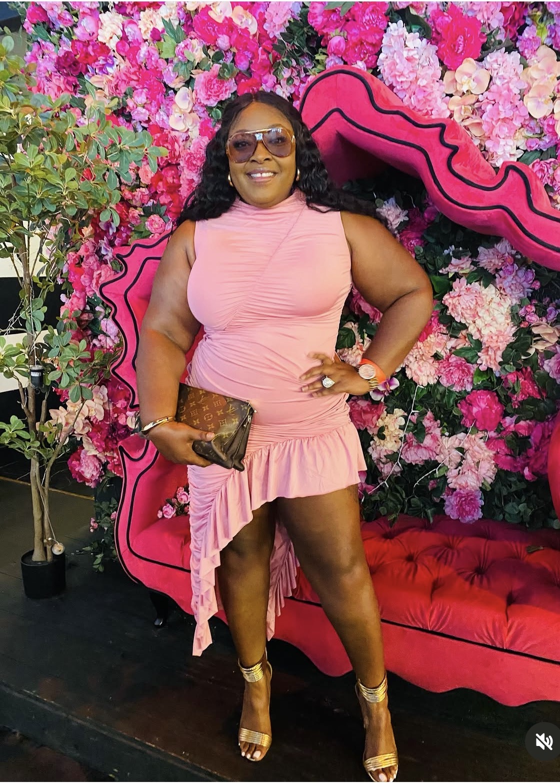 Woman in pink dress with sunglasses, holding a clutch, standing in front of a floral backdrop and pink sofa.