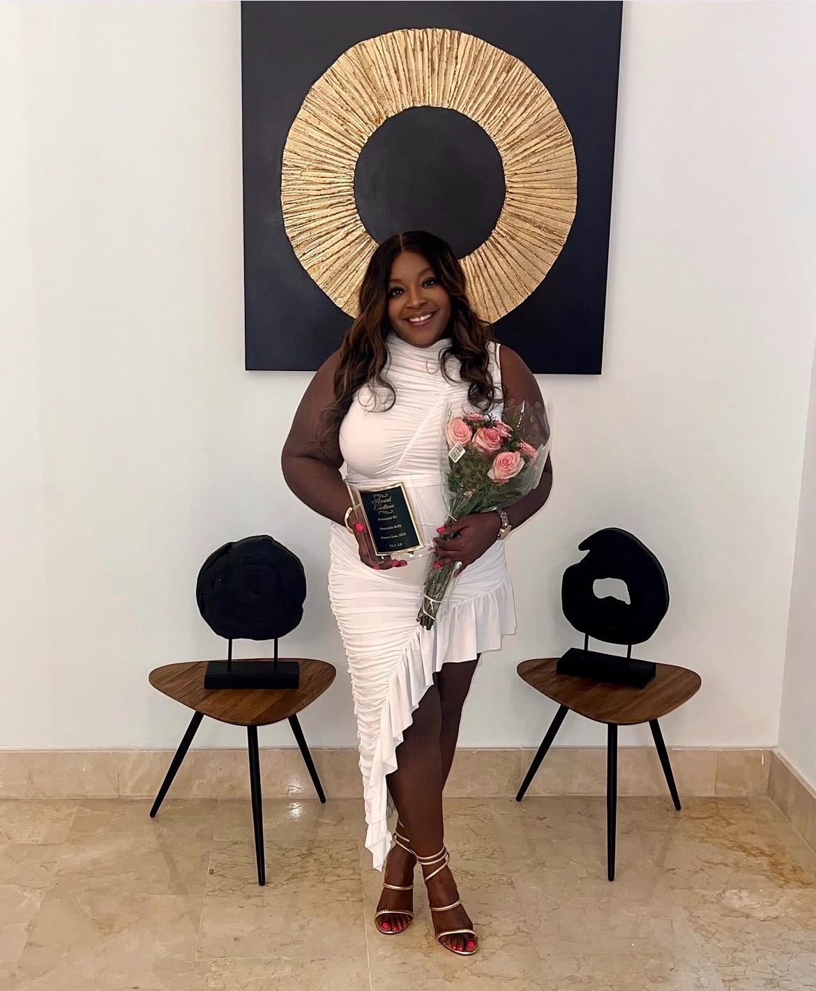 Woman holding a bouquet of pink roses and a phone, standing in front of wall art, smiling, wearing a white dress and heels.