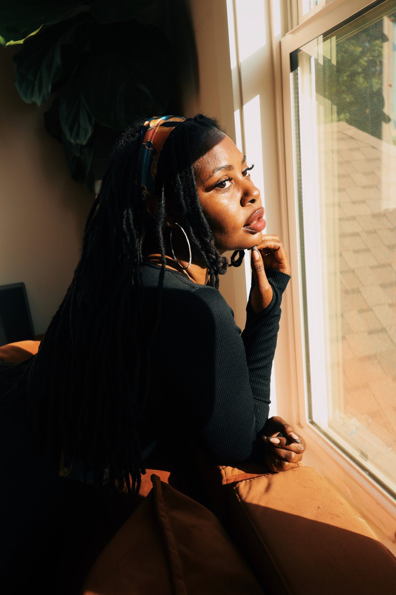 Woman with long hair looking out a window, resting her chin on her hand, indoors with sunlight streaming in.