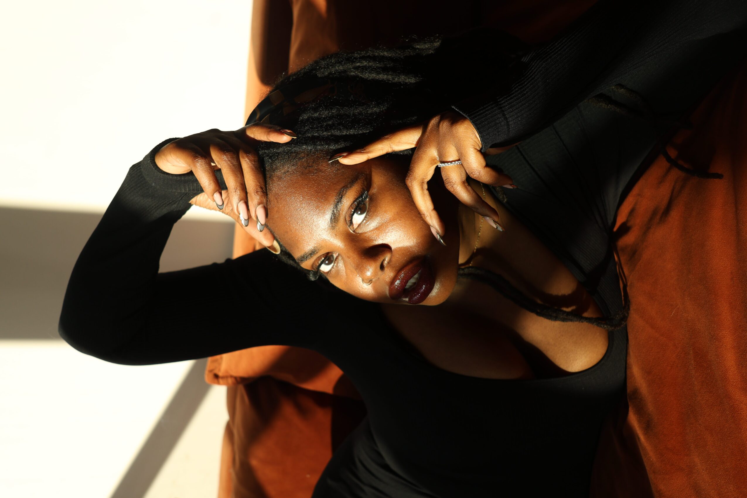 Woman with dark lipstick and makeup, lying on a brown surface, looking at the camera, with her hand near her face.