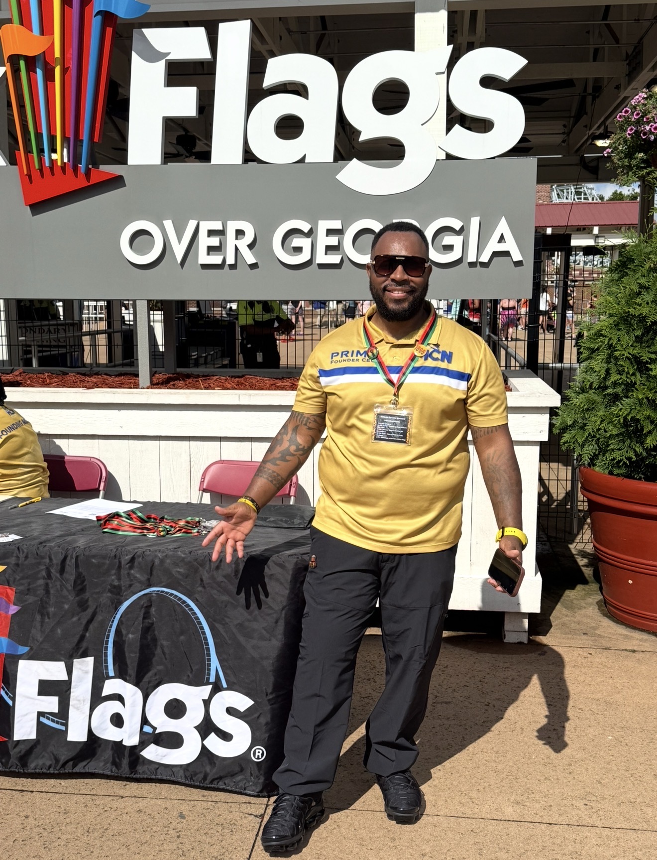 Man in yellow shirt with sunglasses standing outdoors in front of a Flags booth, smiling, with a large Flags sign above.