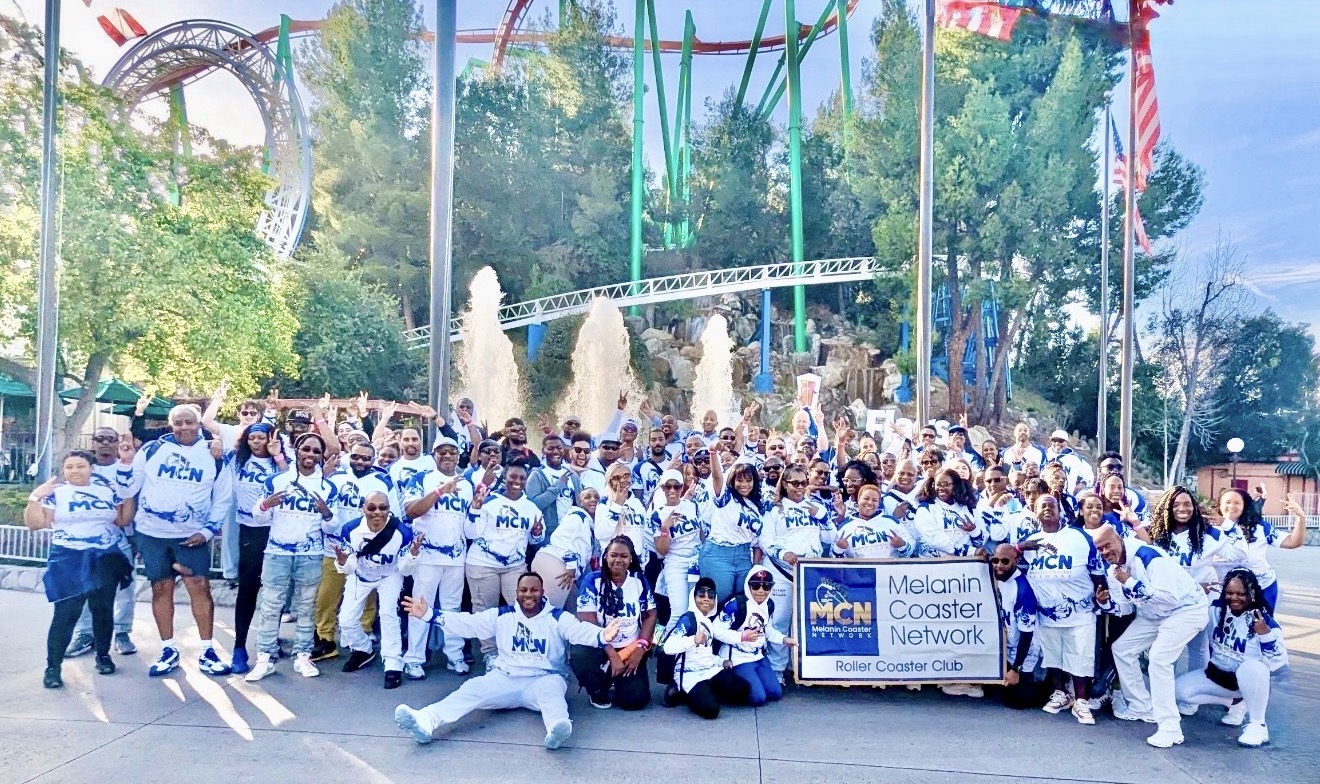 Large group of people in white clothing posing outdoors with amusement park rides and fountains in background.