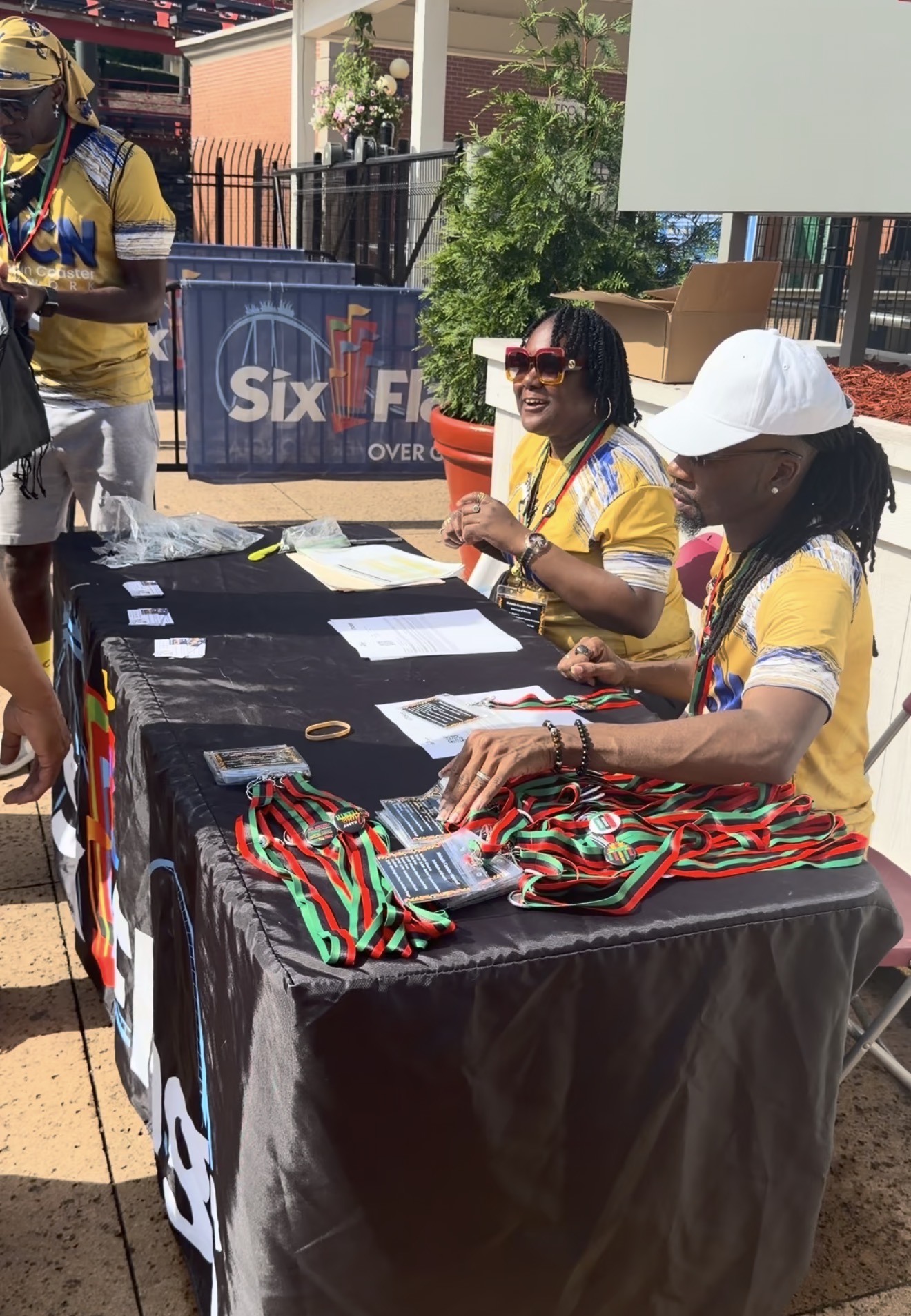 Two women sit at a table with colorful items, outdoors on a sunny day, with people and a banner in the background.