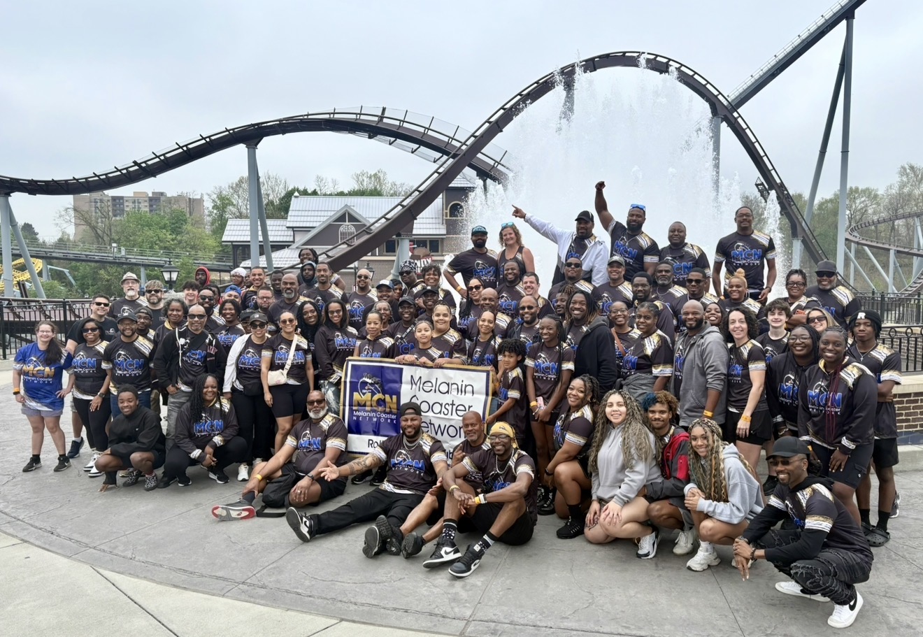 Group of people posing in front of a water fountain at an amusement park, some holding a banner, with a roller coaster in the background.