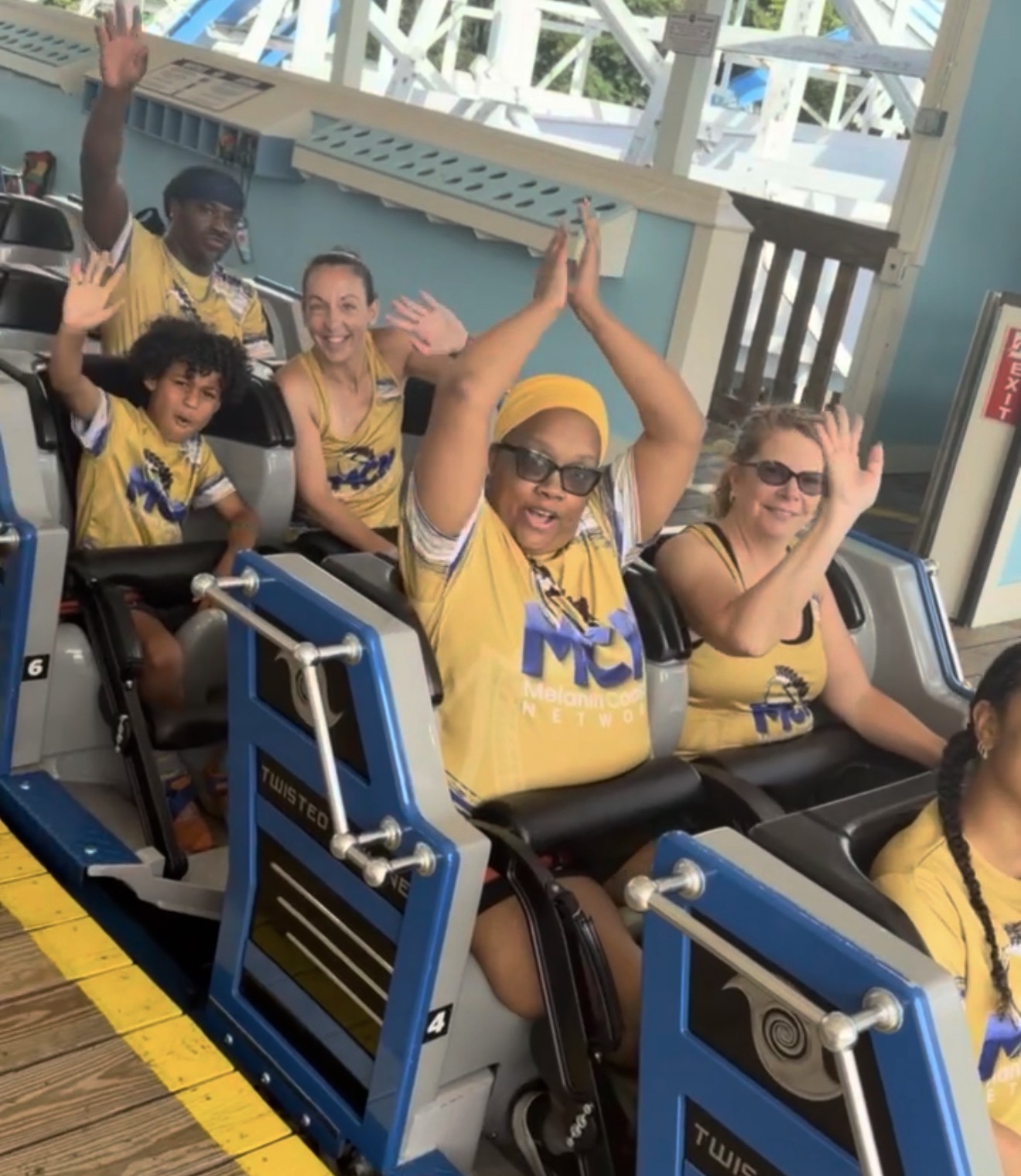 Group of people on a roller coaster ride, smiling and waving, wearing yellow shirts, in an amusement park setting.
