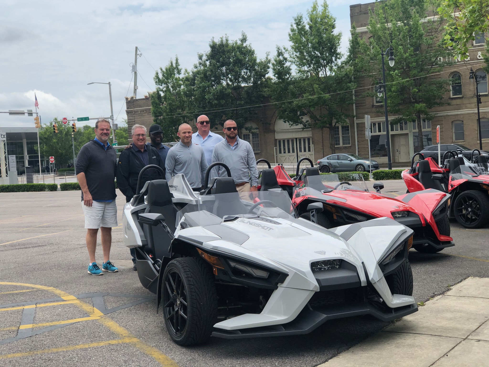 Four people standing next to a white and black three-wheeled vehicle and two red vehicles parked on the street, with trees and buildings in the background.