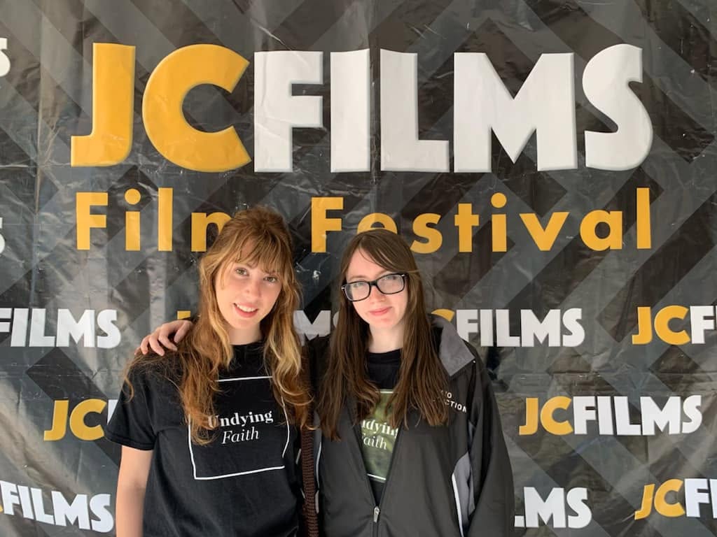 Two young women stand in front of a JCFILMS Film Festival backdrop, smiling and posing for the photo.