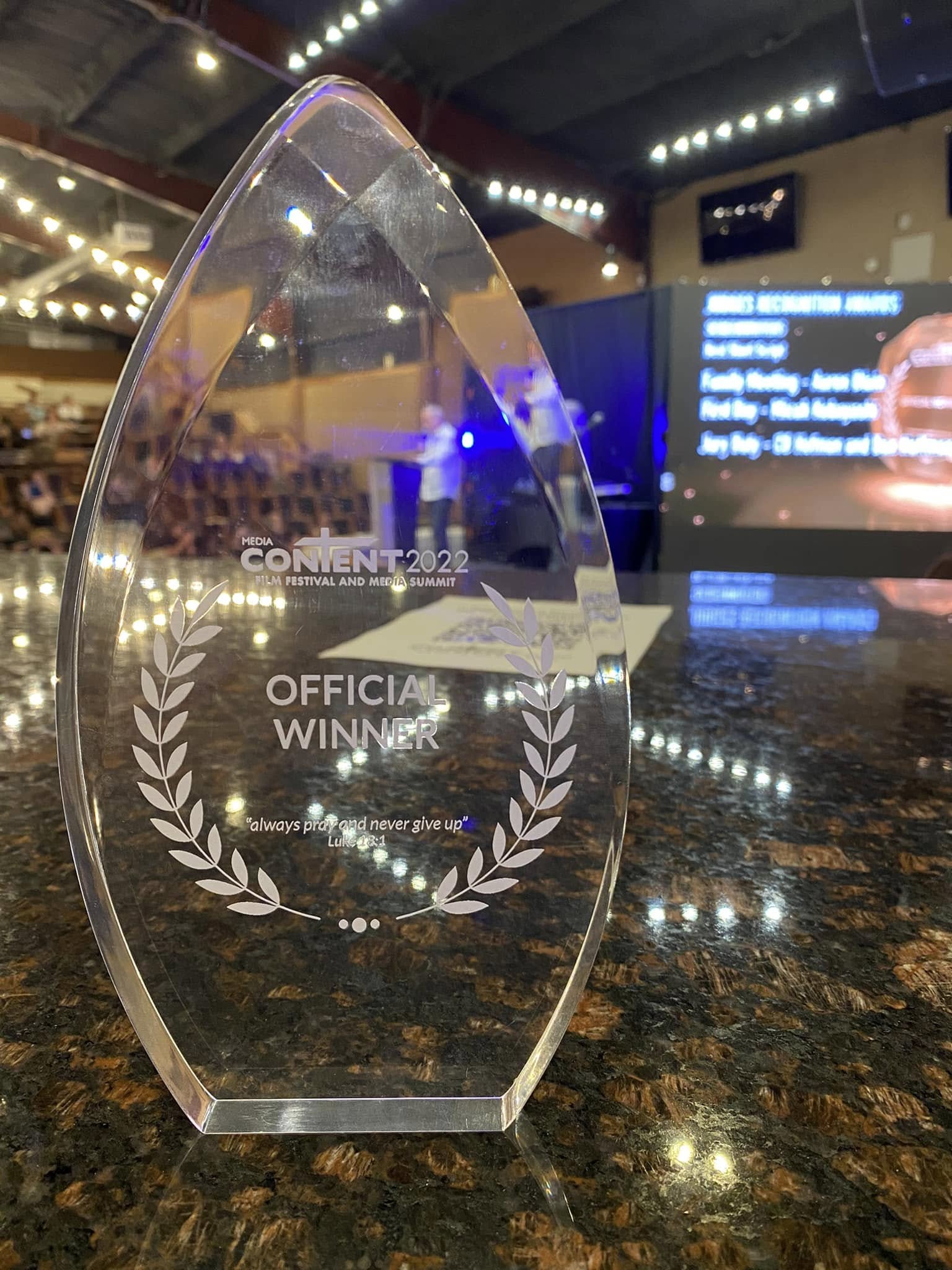 Clear glass award with engraved text and laurel design, placed on a reflective surface in a dimly lit room.