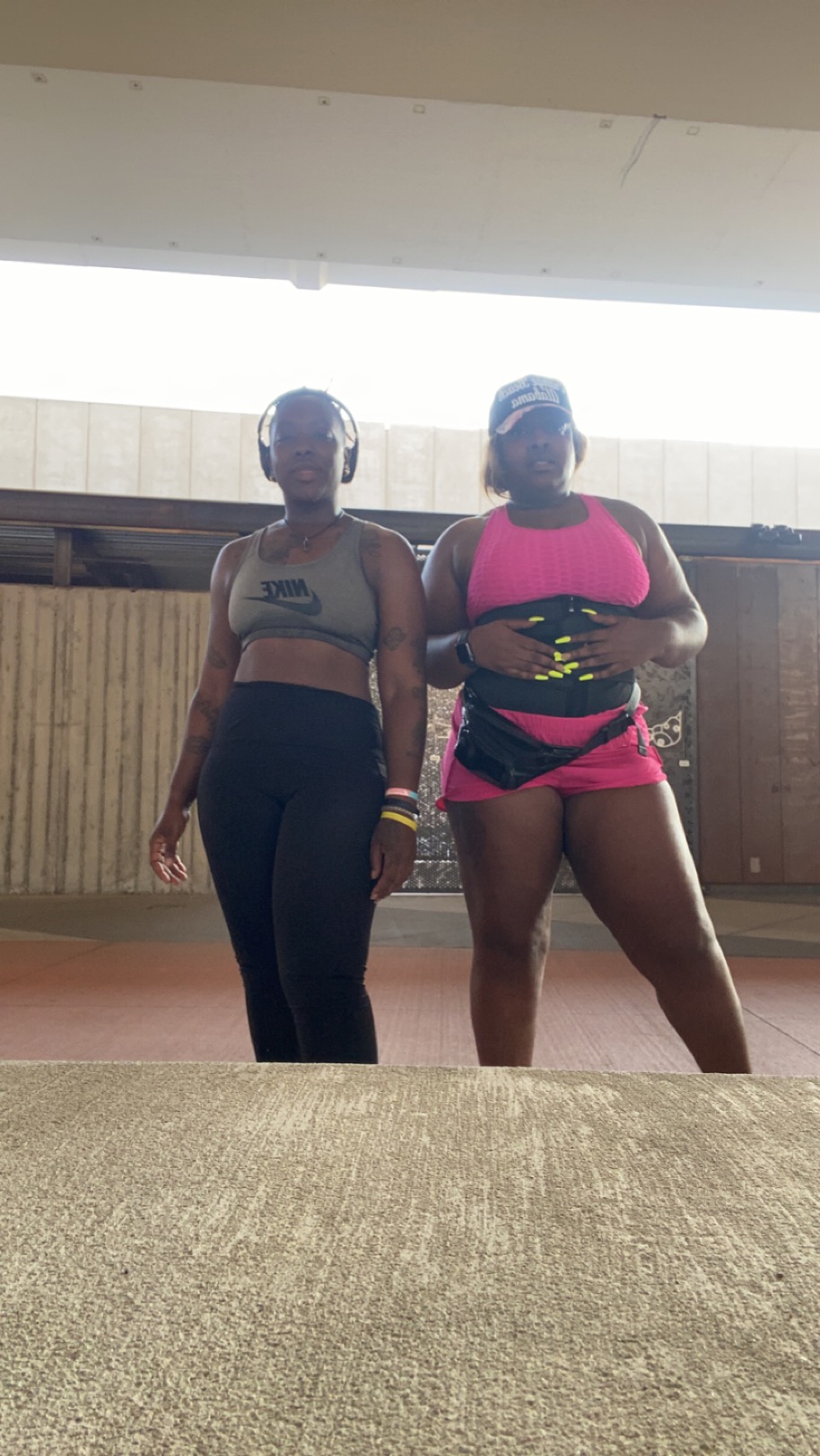 Two women in athletic clothing standing indoors, one in black leggings and the other in pink shorts and a pink top, both wearing helmets.